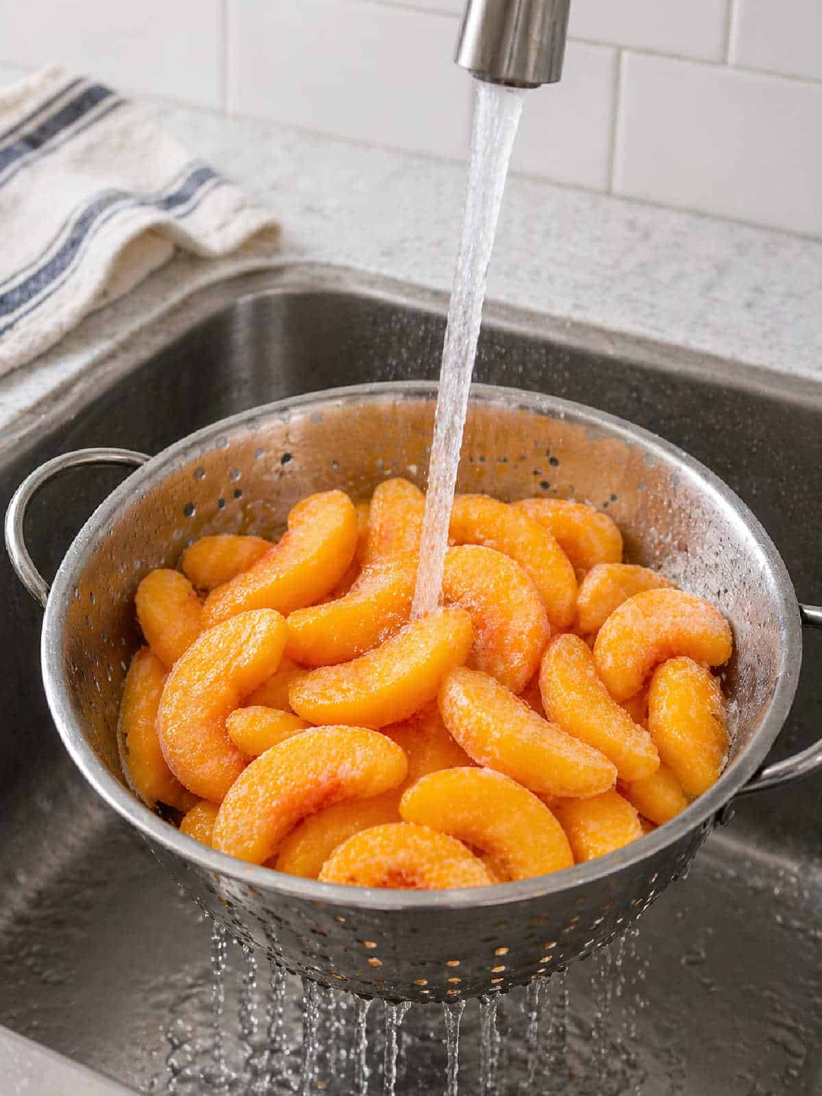 A stream of water rinses frozen peach slices in a metal colander placed in a kitchen sink.
