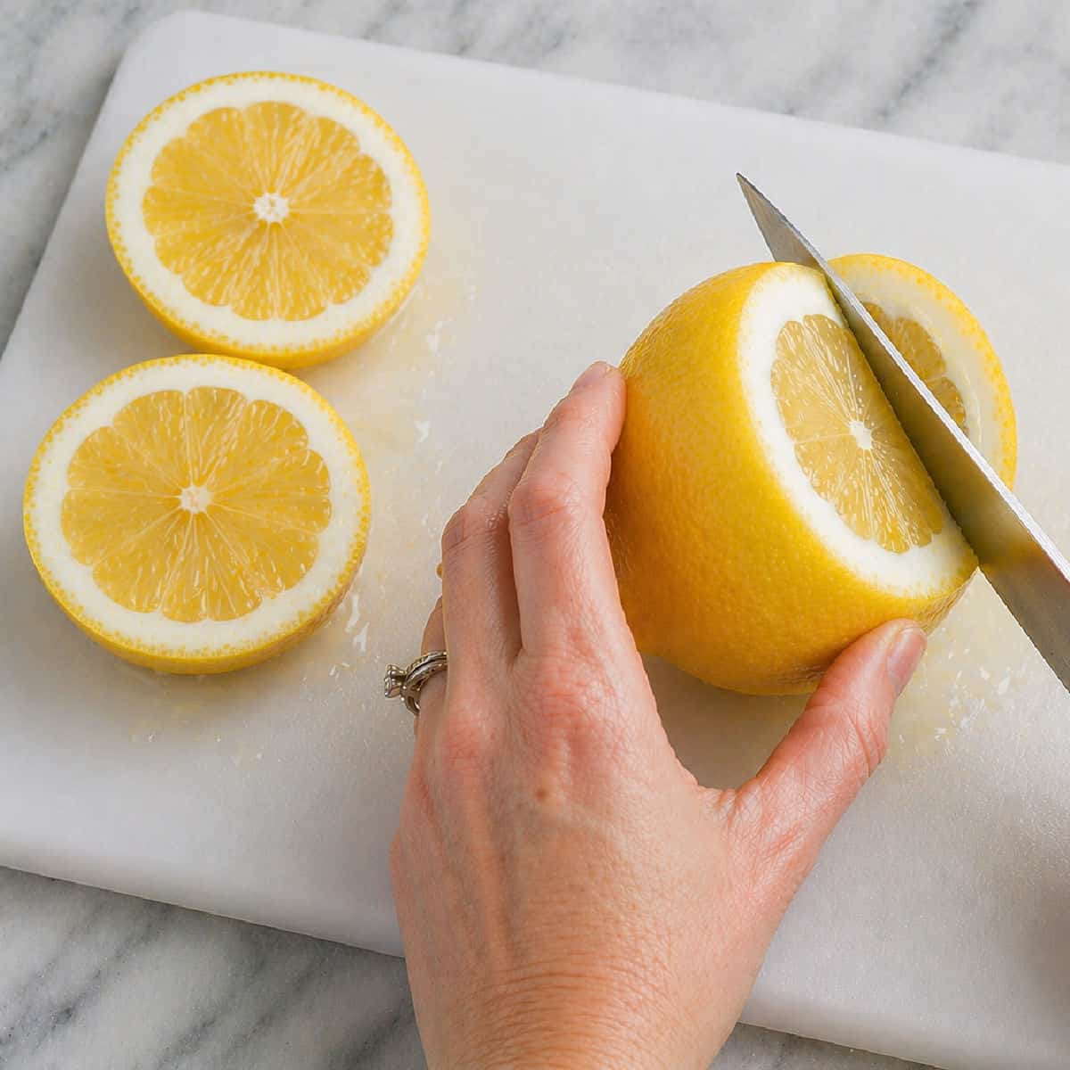 A hand holding a lemon while slicing it on a white cutting board with two lemon halves beside it.