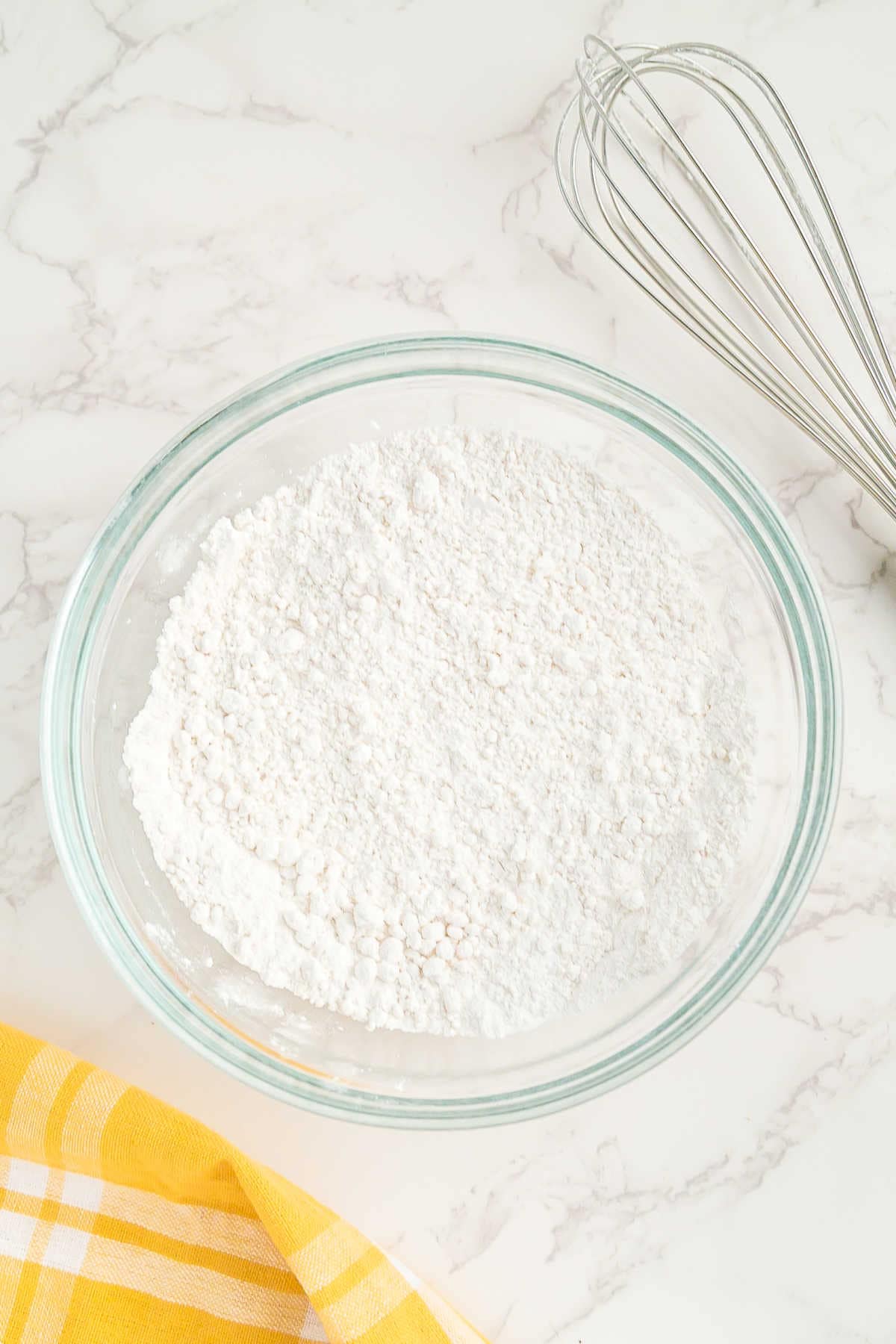 A glass bowl filled with flour mixture on a marble countertop, with a metal whisk and a yellow checkered cloth nearby.