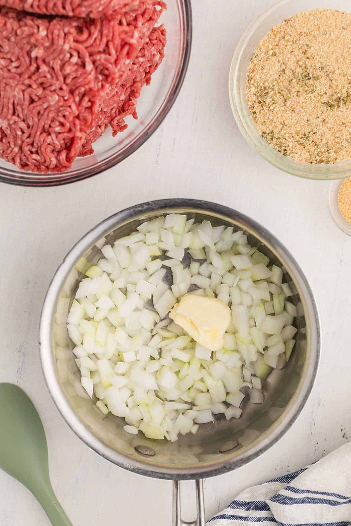 Chopped onions and a pat of butter in a pot, with raw ground beef and breadcrumbs in bowls nearby on a white surface.