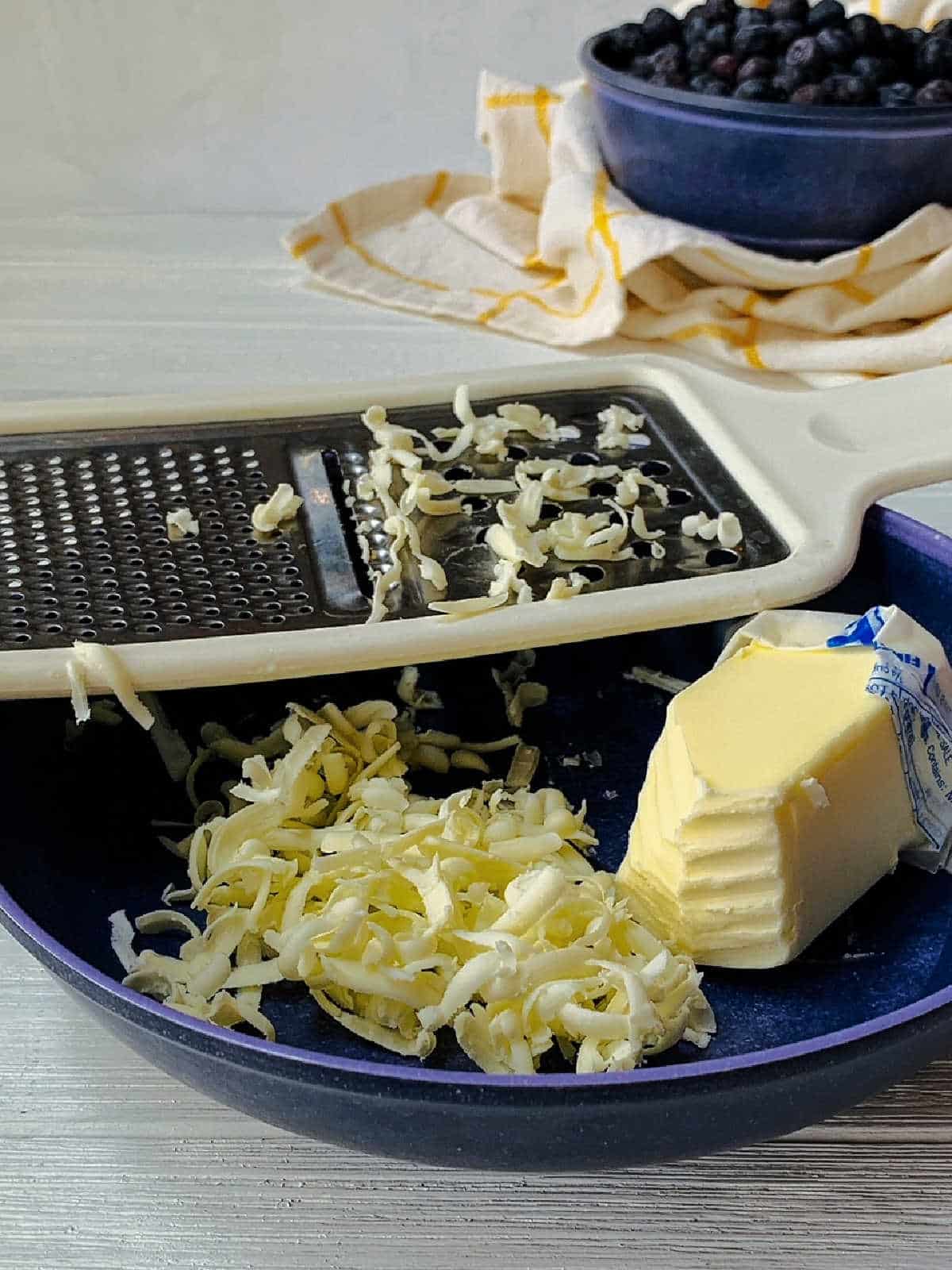 A cheese grater with shredded butter, a partially unwrapped stick of butter, and a blue bowl of blueberries in the background.