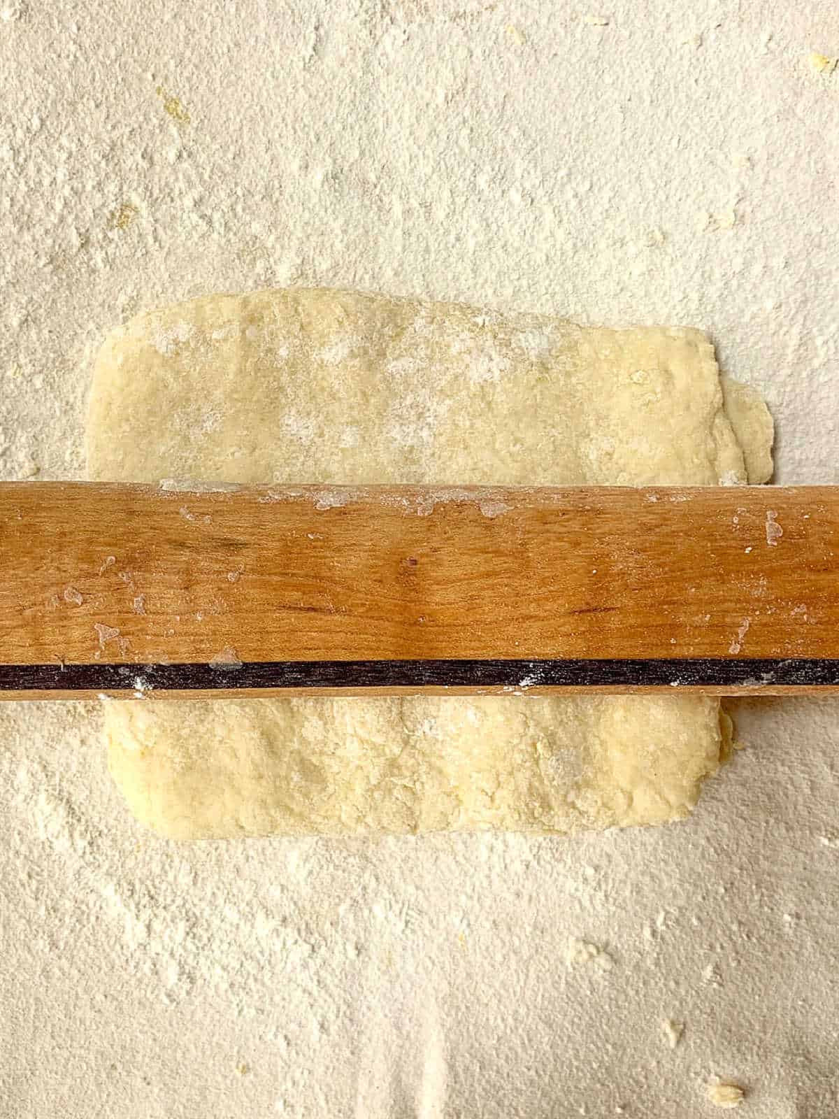 A wooden rolling pin presses a rectangular piece of dough on a floured surface.