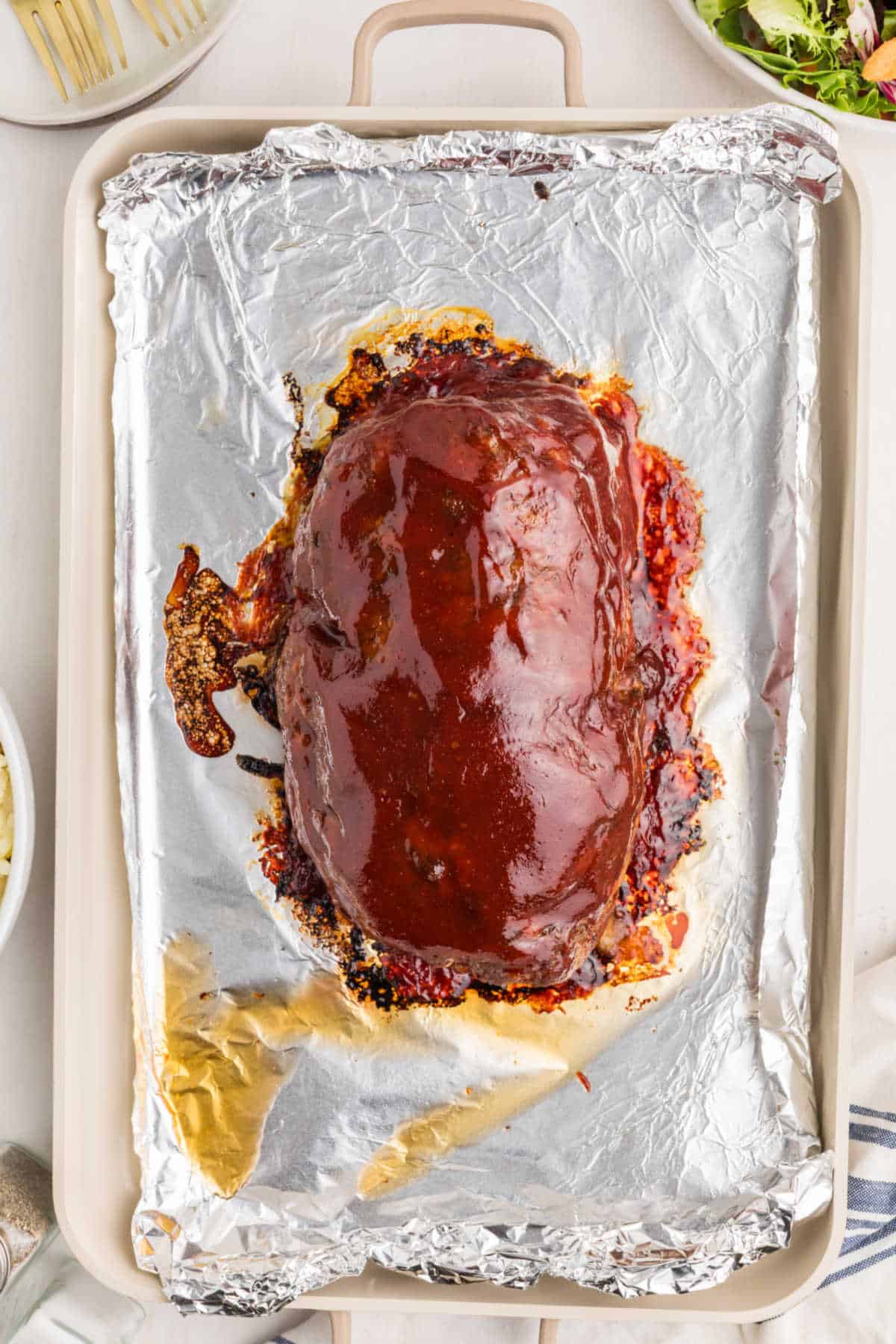 A baked meatloaf covered in a shiny glaze sits on a foil-lined baking sheet, surrounded by side dishes on a white table.