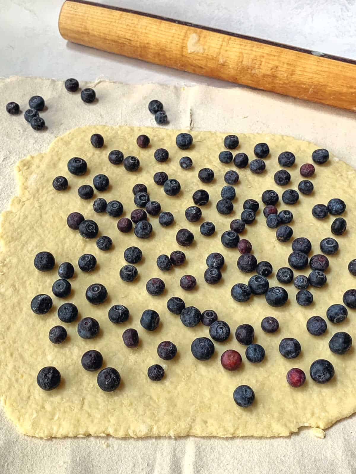 Rolled out dough topped with scattered fresh blueberries, with a wooden rolling pin in the background on a light surface.