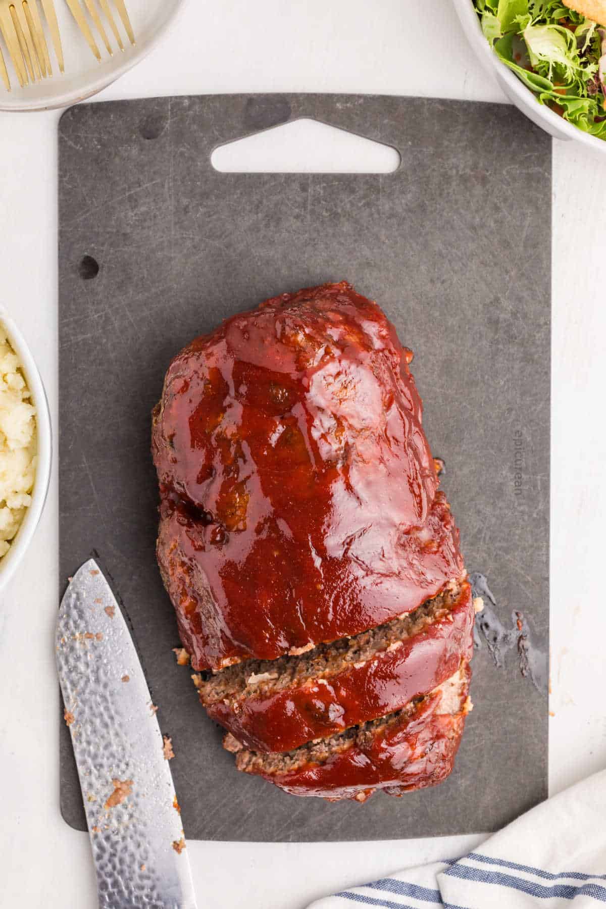 A glazed meatloaf with two slices partially cut sits on a dark cutting board; a knife and side dishes are visible nearby.