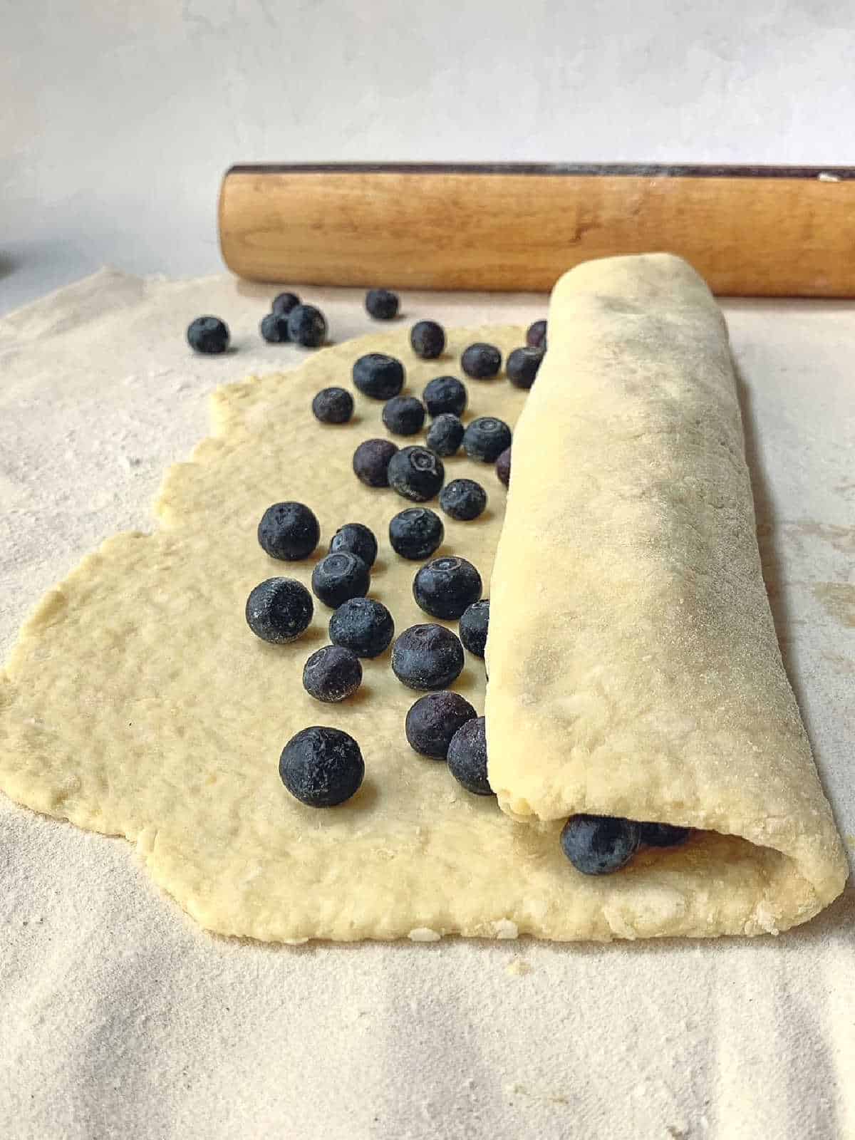 Unbaked dough being rolled up with fresh blueberries on a floured surface, with a wooden rolling pin in the background.