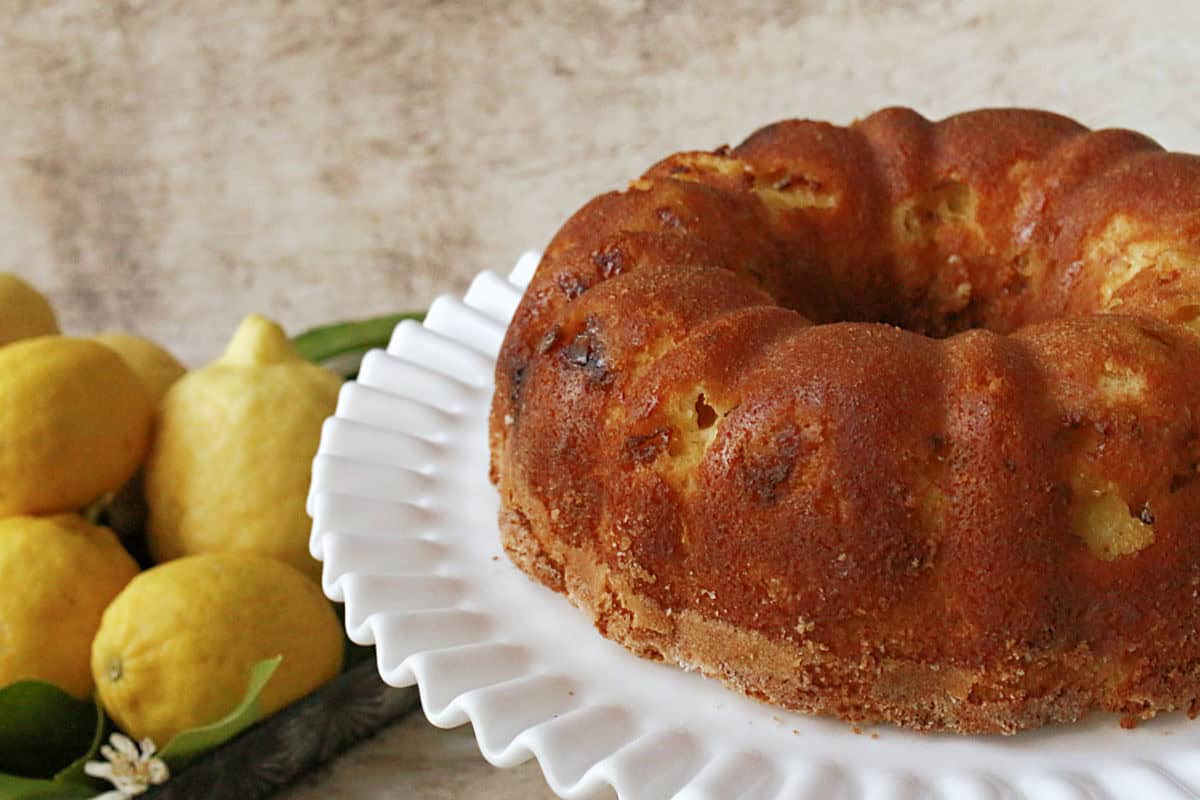 A golden-brown bundt cake on a white scalloped cake stand, with fresh lemons and green leaves in the background.
