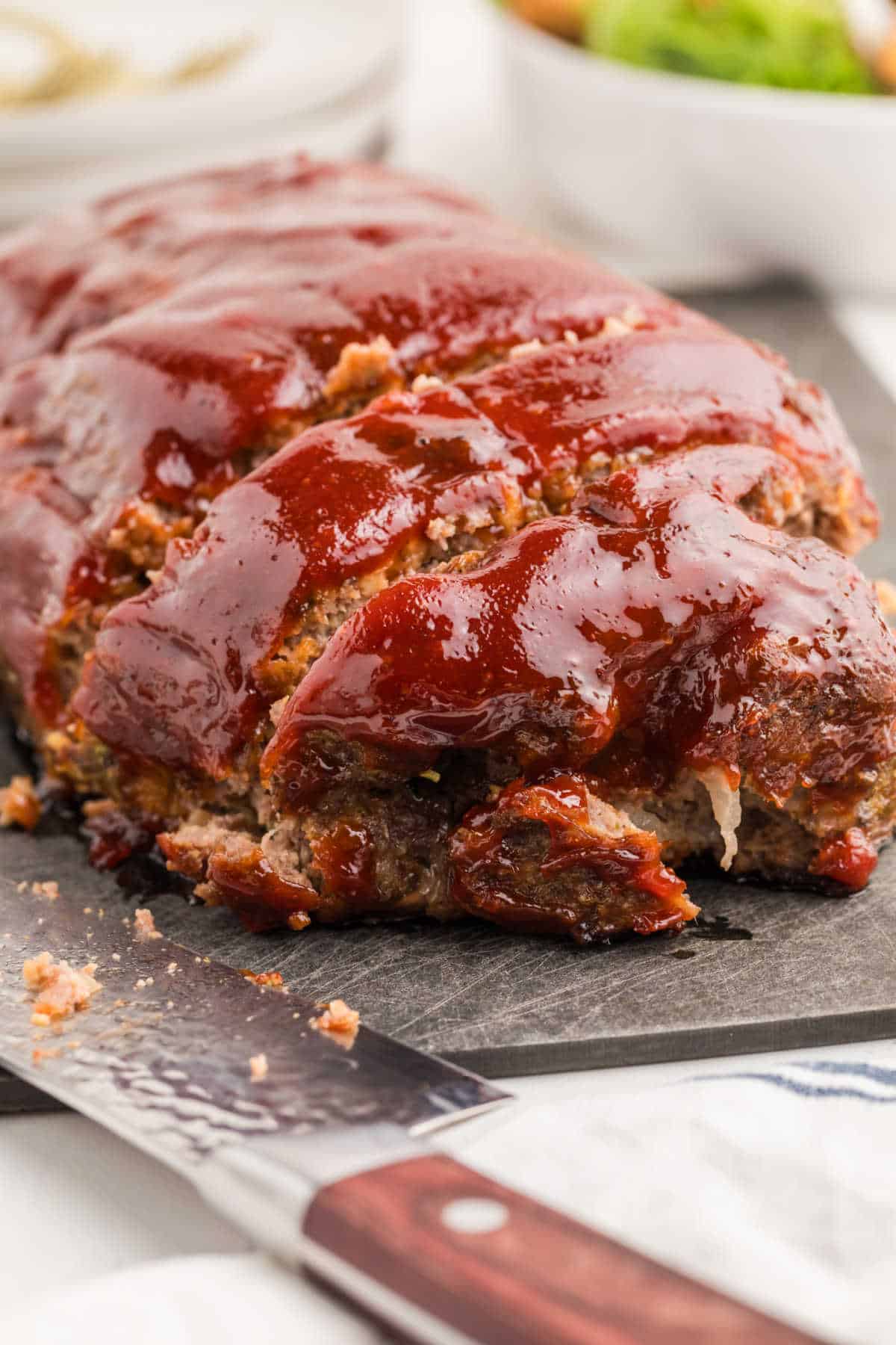 A sliced meatloaf topped with a glossy layer of ketchup glaze sits on a cutting board next to a knife.
