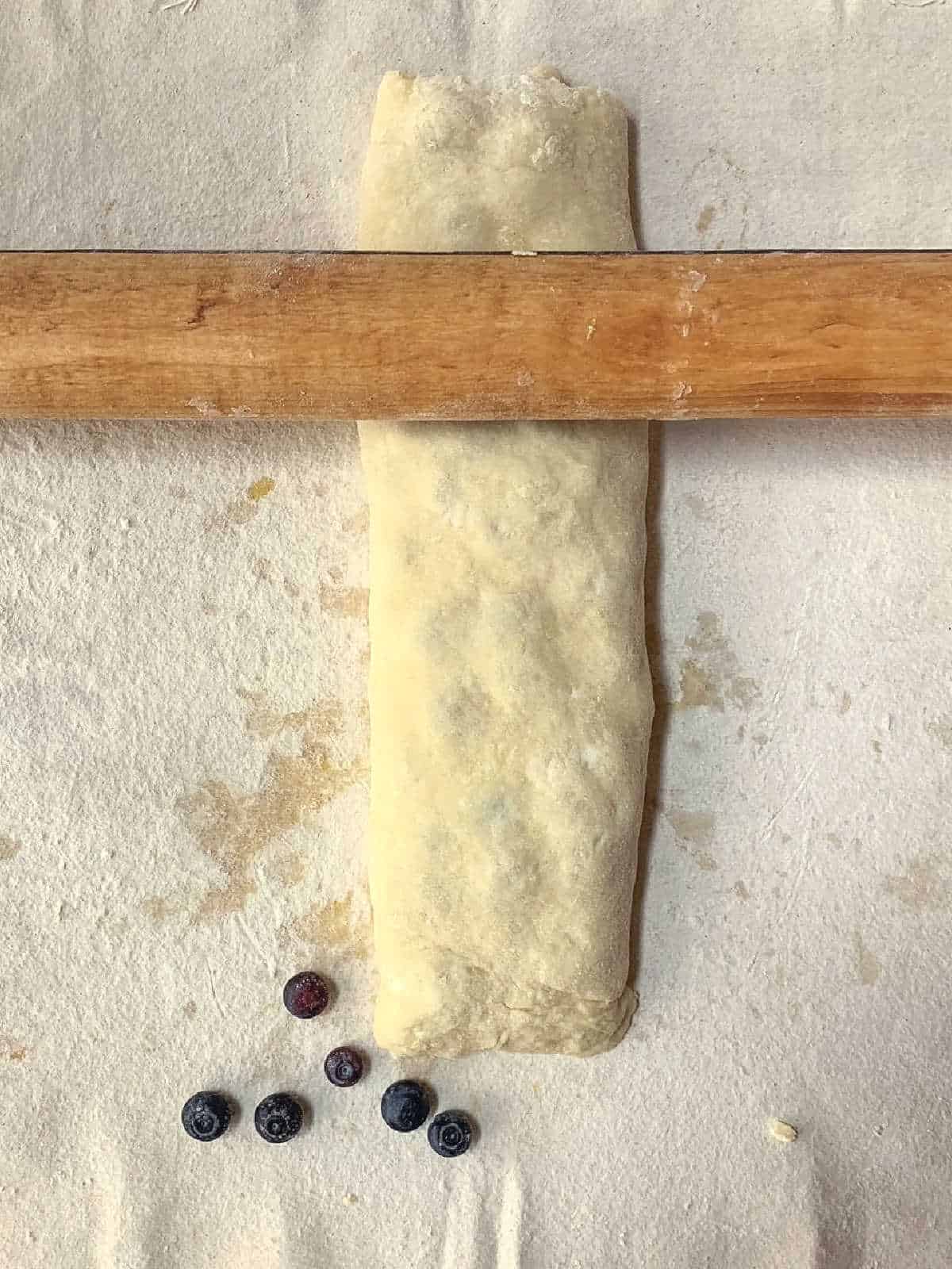 A rolled dough log on a floured surface with a wooden rolling pin above it and several blueberries scattered nearby.