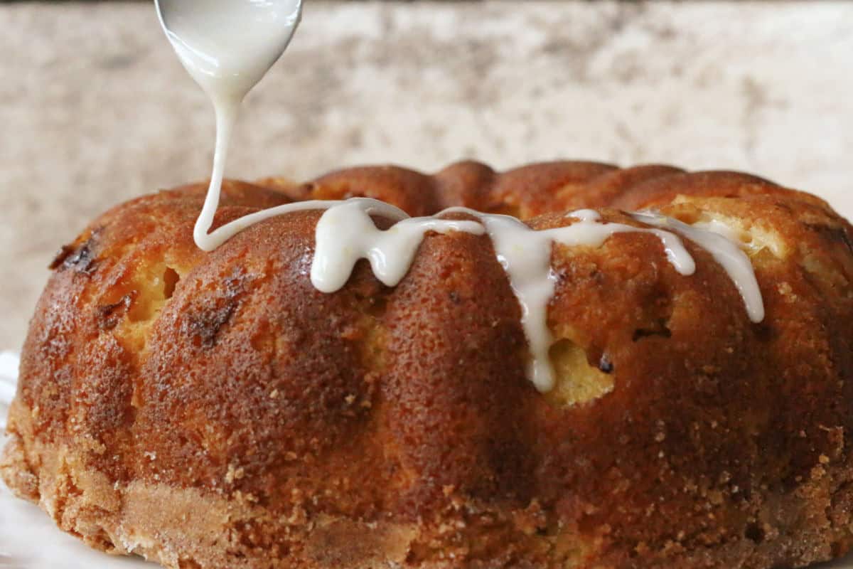 A spoon drizzles white icing over a round, golden-brown bundt cake on a neutral background.