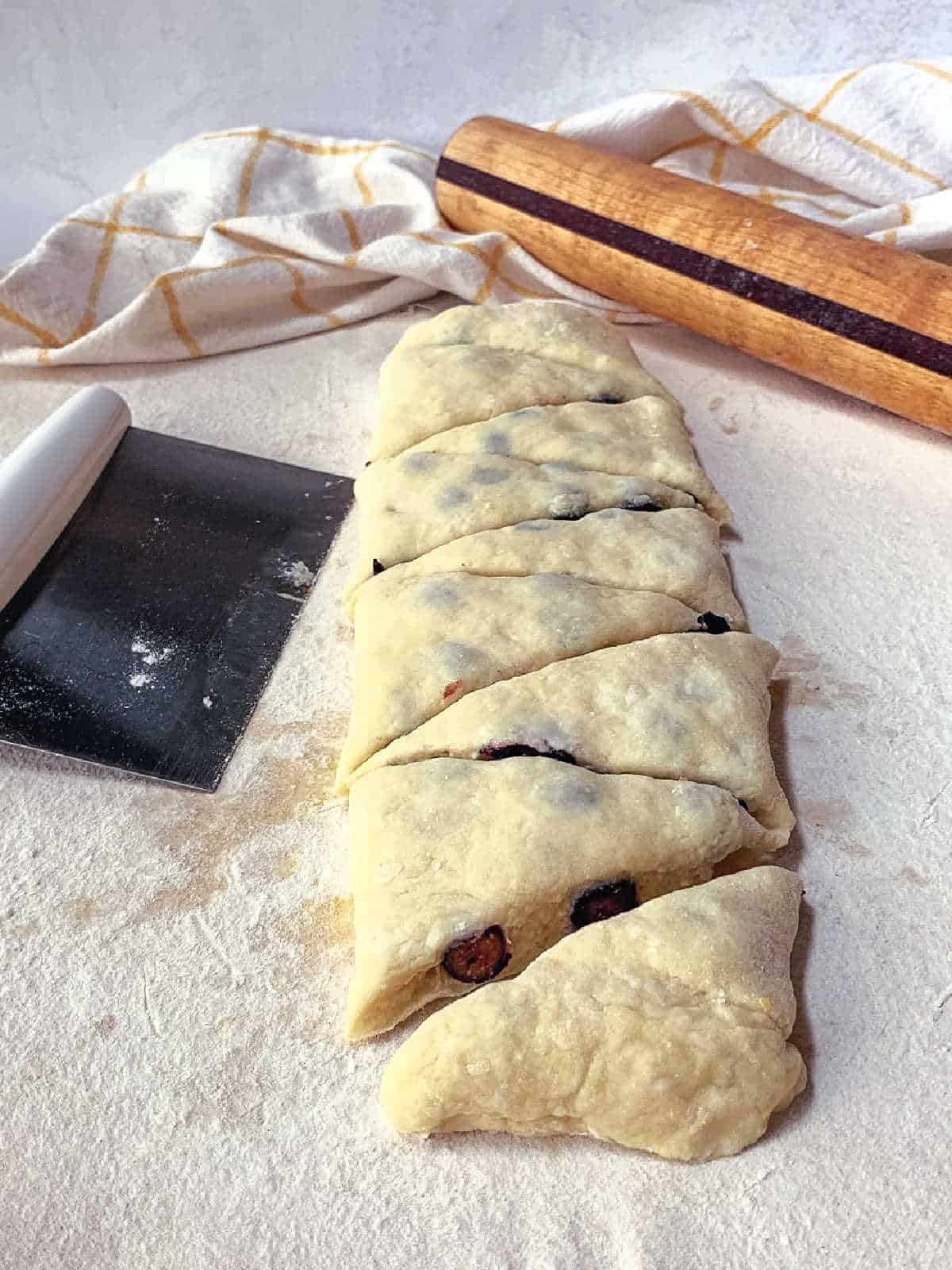 Sliced, unbaked scone dough with visible berries rests on a floured surface next to a dough scraper, rolling pin, and dish towel.