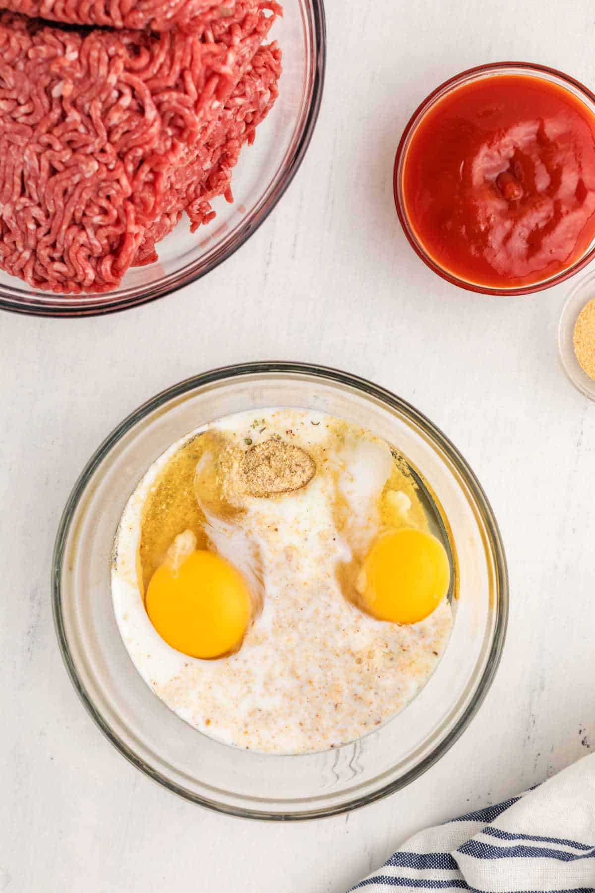 Glass bowls with ground beef, ketchup, and a mixture of eggs, milk, breadcrumbs, and seasonings on a white surface.