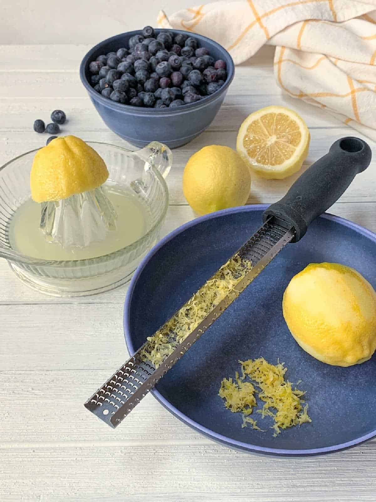 A zested lemon, microplane grater, lemon zest, two whole lemons, a juicer with half a lemon, and a bowl of blueberries on a white surface.