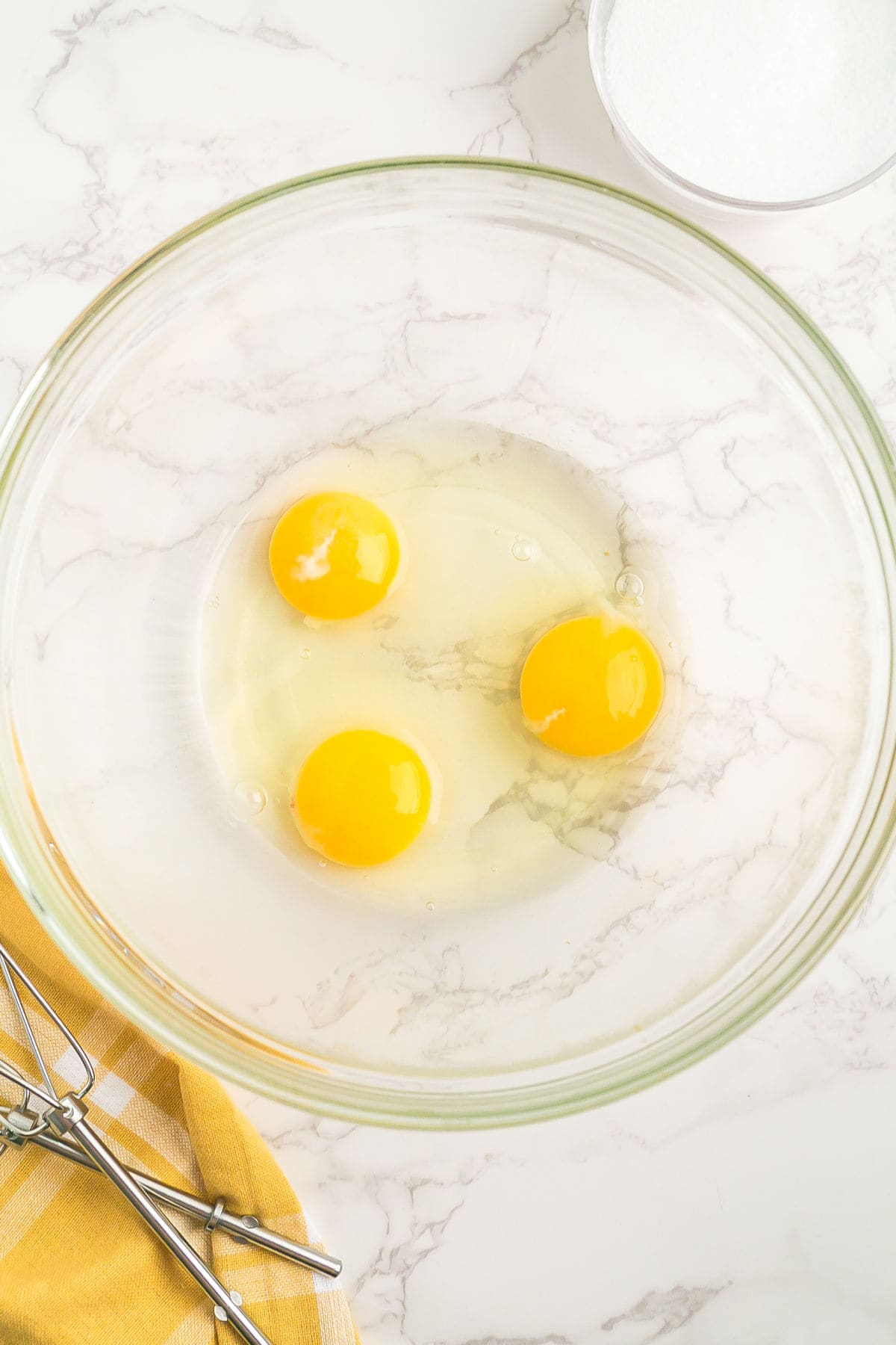 Three raw eggs in a clear glass mixing bowl on a marble countertop, with a yellow towel, metal beaters, and a bowl of sugar nearby.