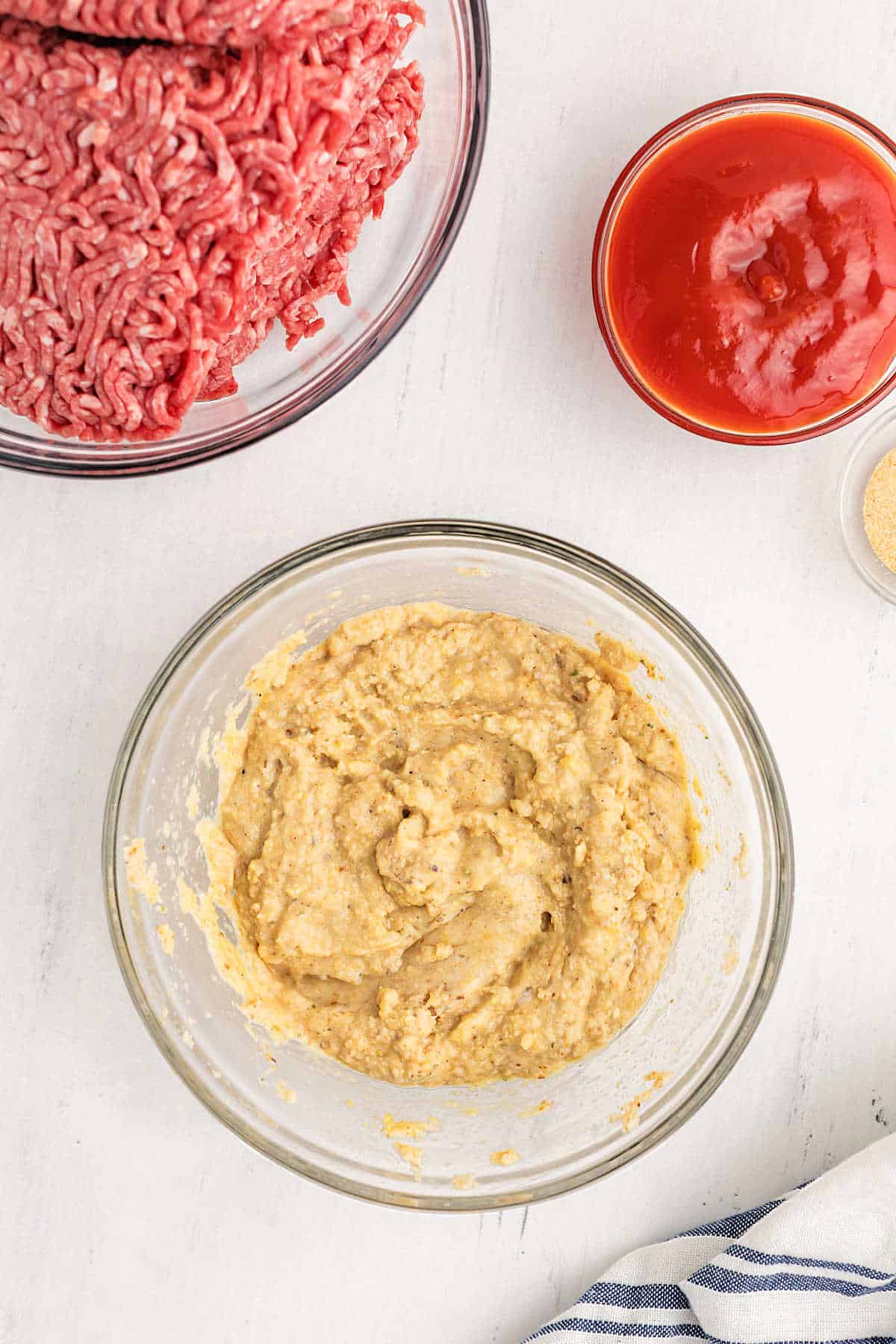 A bowl of ground beef, a bowl of ketchup, and a bowl of a beige, mixed batter sit on a white surface with a striped towel partially visible.