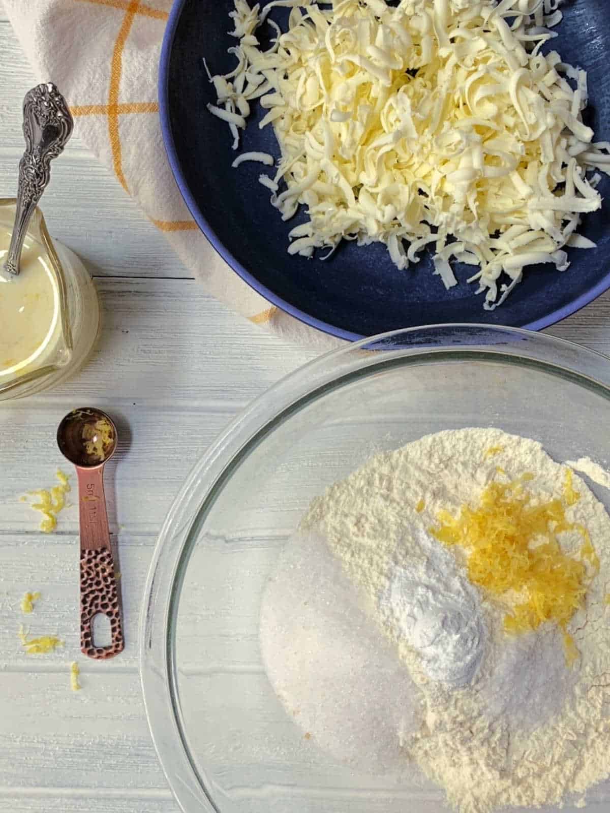 A glass bowl with flour, sugar, baking powder, and lemon zest, next to a bowl of shredded cheese, a jar of white liquid, a towel, and a measuring spoon on a white surface.