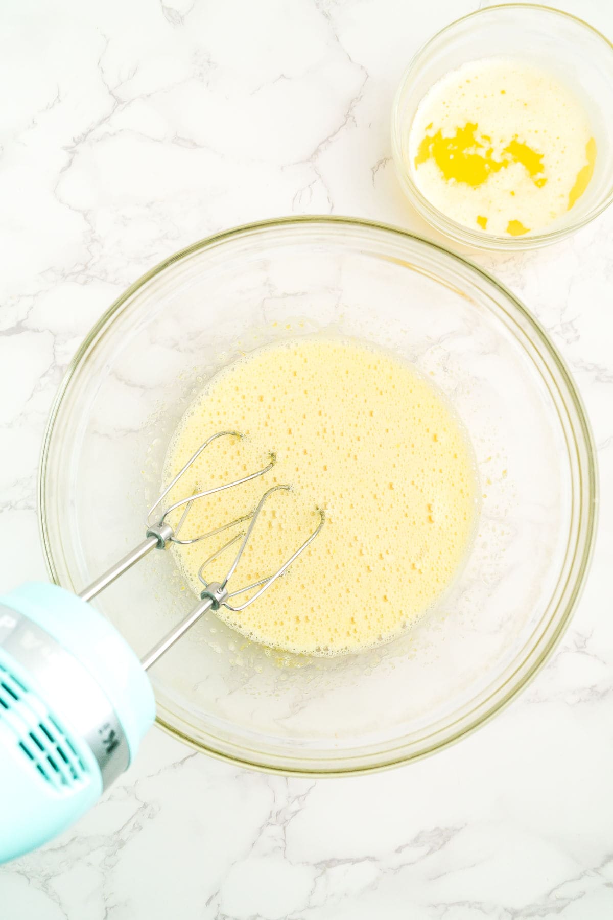 A hand mixer with beaters is mixing a yellow batter in a glass bowl on a marble surface, with another bowl of melted butter nearby.
