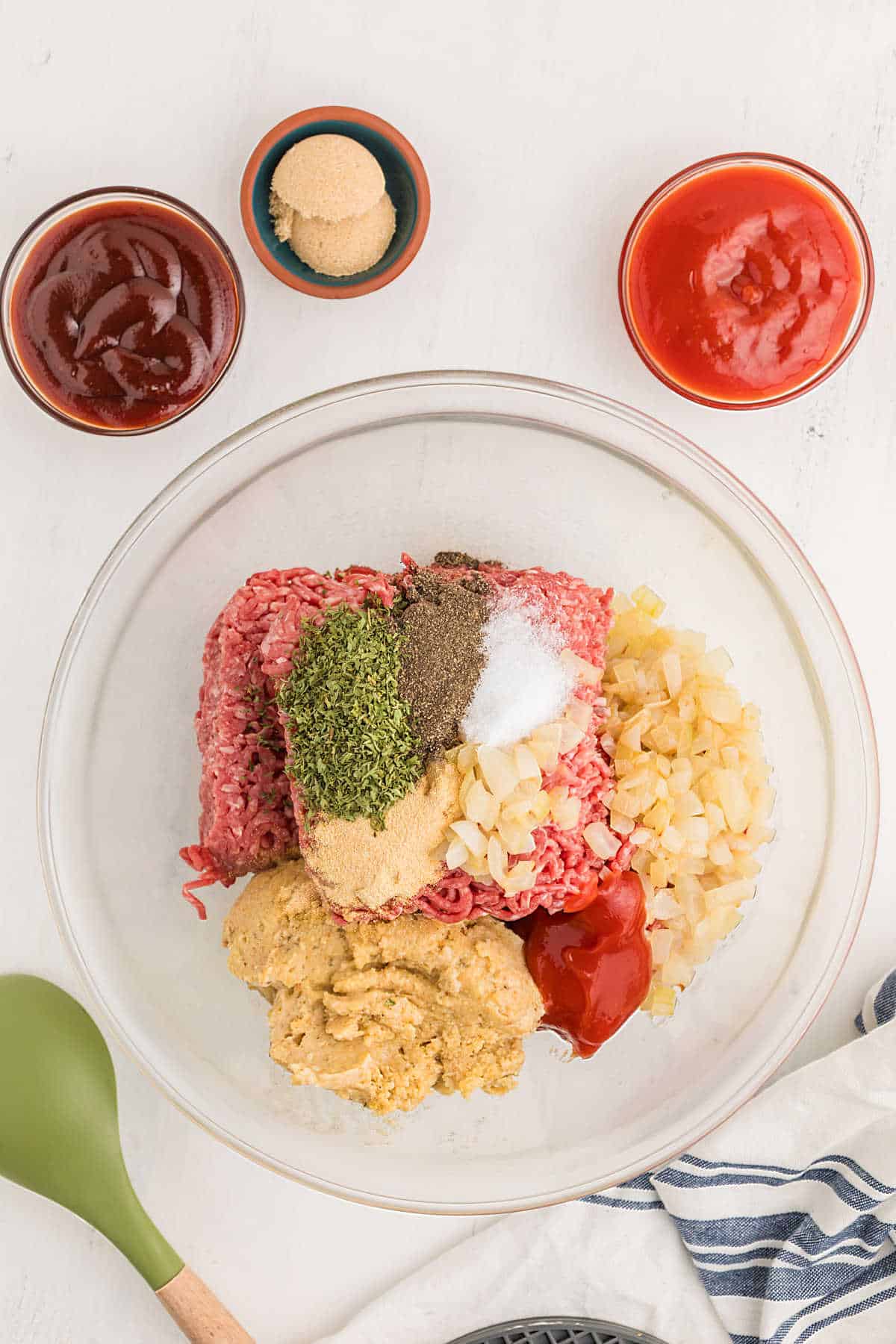 A glass bowl contains raw ground beef, chopped onions, seasonings, ketchup, mustard, and spices. Surrounding bowls hold ketchup, barbecue sauce, and brown sugar.