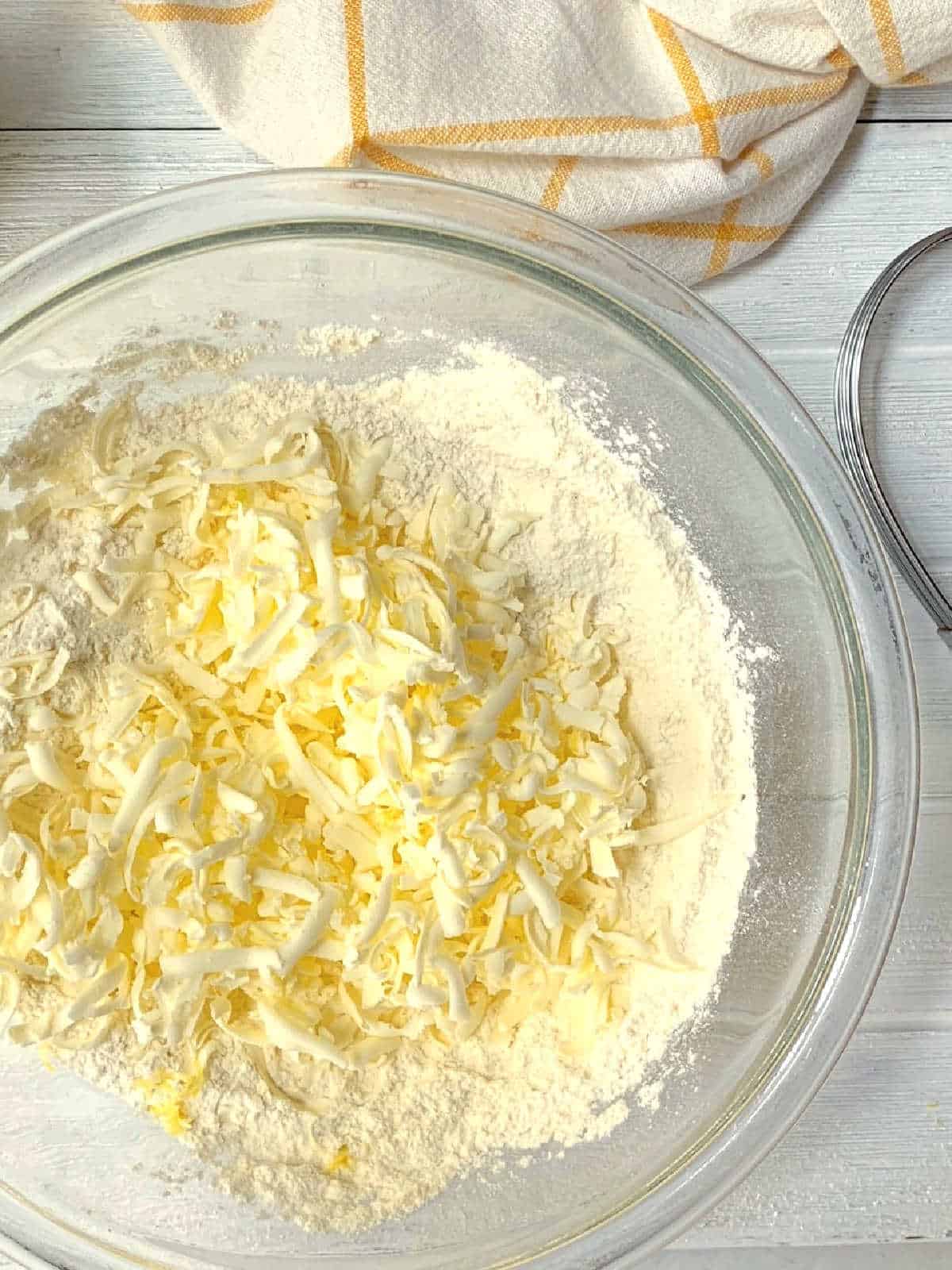 Large glass bowl with flour and shredded butter on a white surface, next to a metal whisk and a white and yellow checkered kitchen towel.