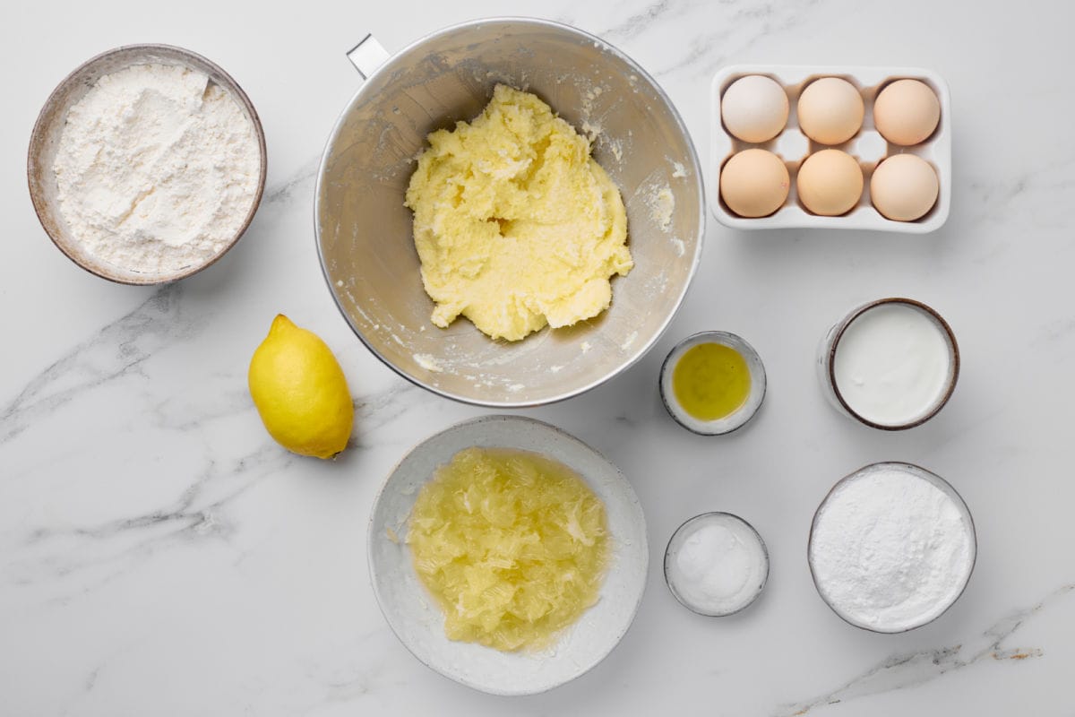 Baking ingredients on a marble surface, including flour, eggs, lemon, creamed butter and sugar in a bowl, oil, yogurt, shredded apple, salt, and powdered sugar.