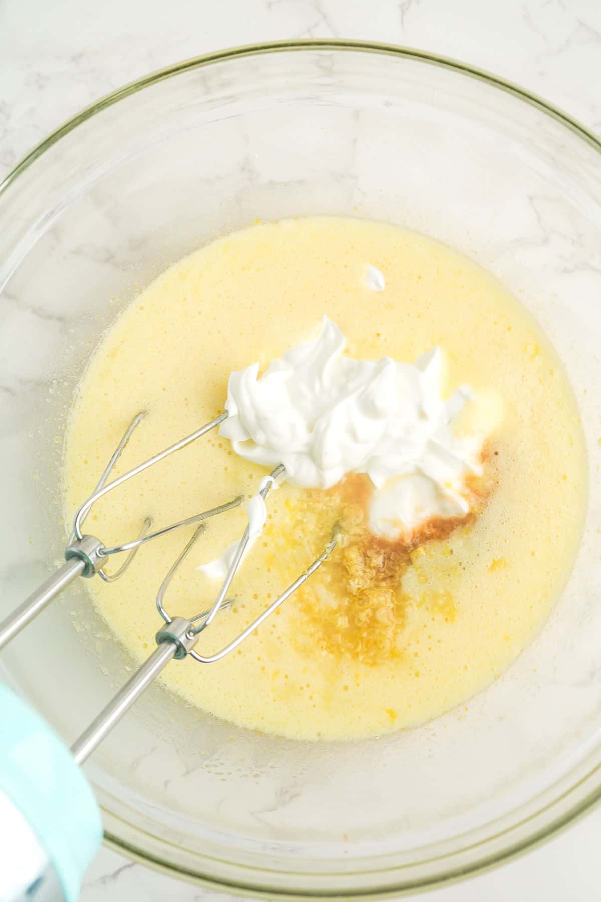 A glass bowl containing a yellow batter mixture with dollops of white yogurt, vanilla extract, and an electric hand mixer on a marble countertop.