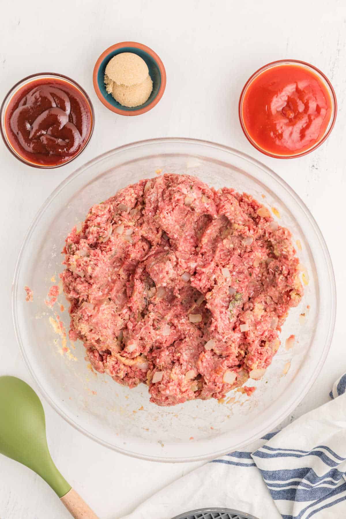 A glass bowl containing mixed raw ground meat with chopped onions, surrounded by small bowls of ketchup, brown sugar, tomato sauce, and a green spatula on a white surface.