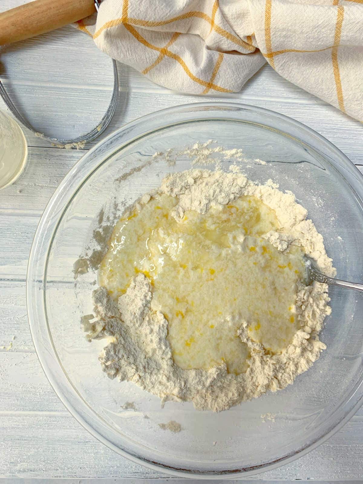 A glass bowl with flour and a liquid mixture being combined, a spoon in the bowl, with a rolling pin and kitchen towel nearby on a white surface.