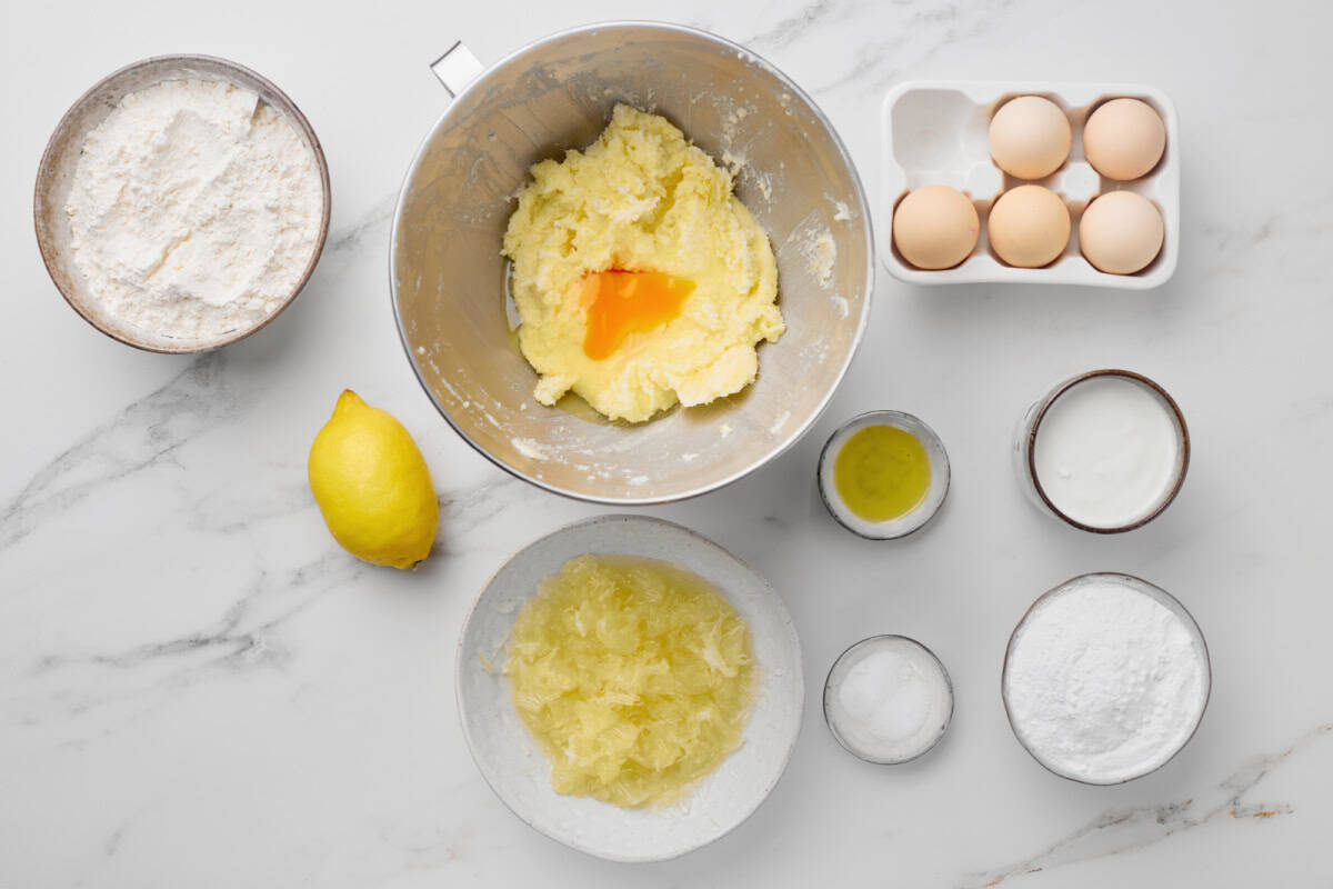 Baking ingredients on a marble surface: flour, eggs, a lemon, creamed butter with an egg, oil, yogurt, sugar, salt, and grated lemon zest in separate bowls.