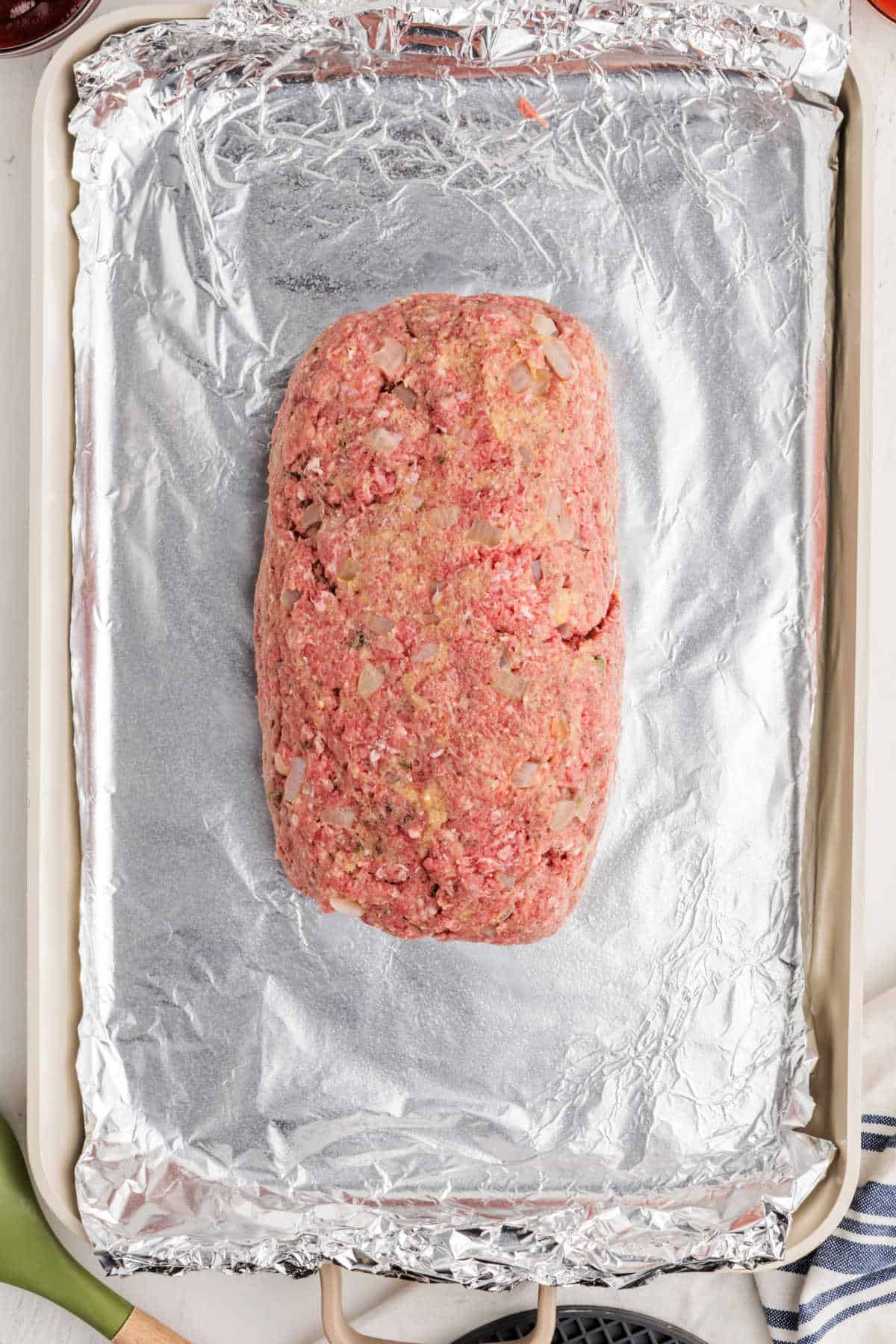 A raw meatloaf mixture shaped into a loaf sits on a foil-lined baking sheet, ready to be cooked.