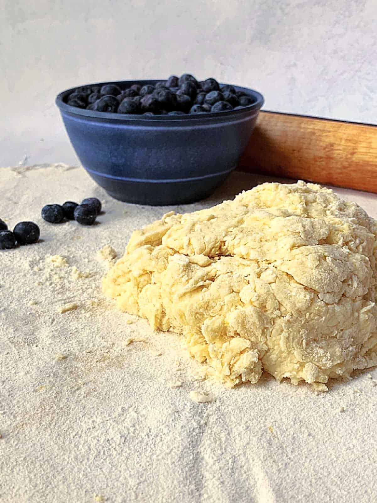 A mound of raw scone dough sits on a floured surface with a blue bowl of blueberries and a rolling pin in the background.