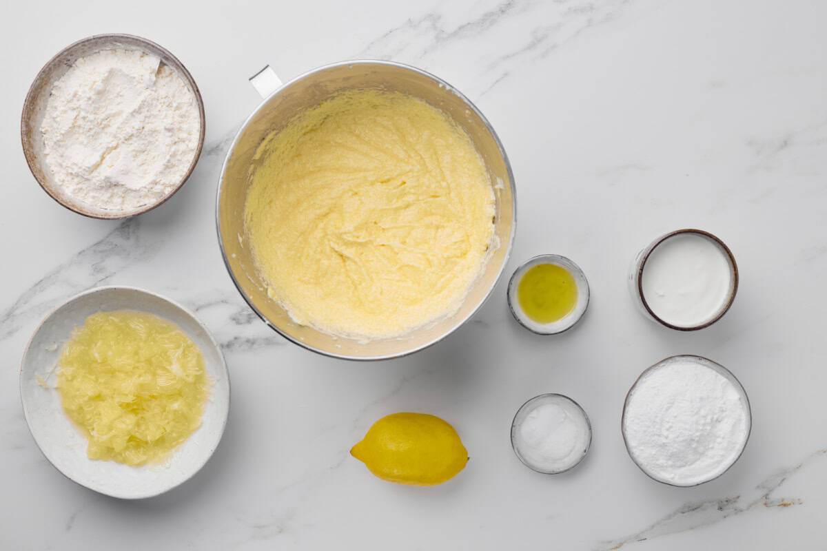 Baking ingredients arranged on a white surface, including flour, sugar, lemon, lemon zest, oil, yogurt, and a mixing bowl with creamed batter.