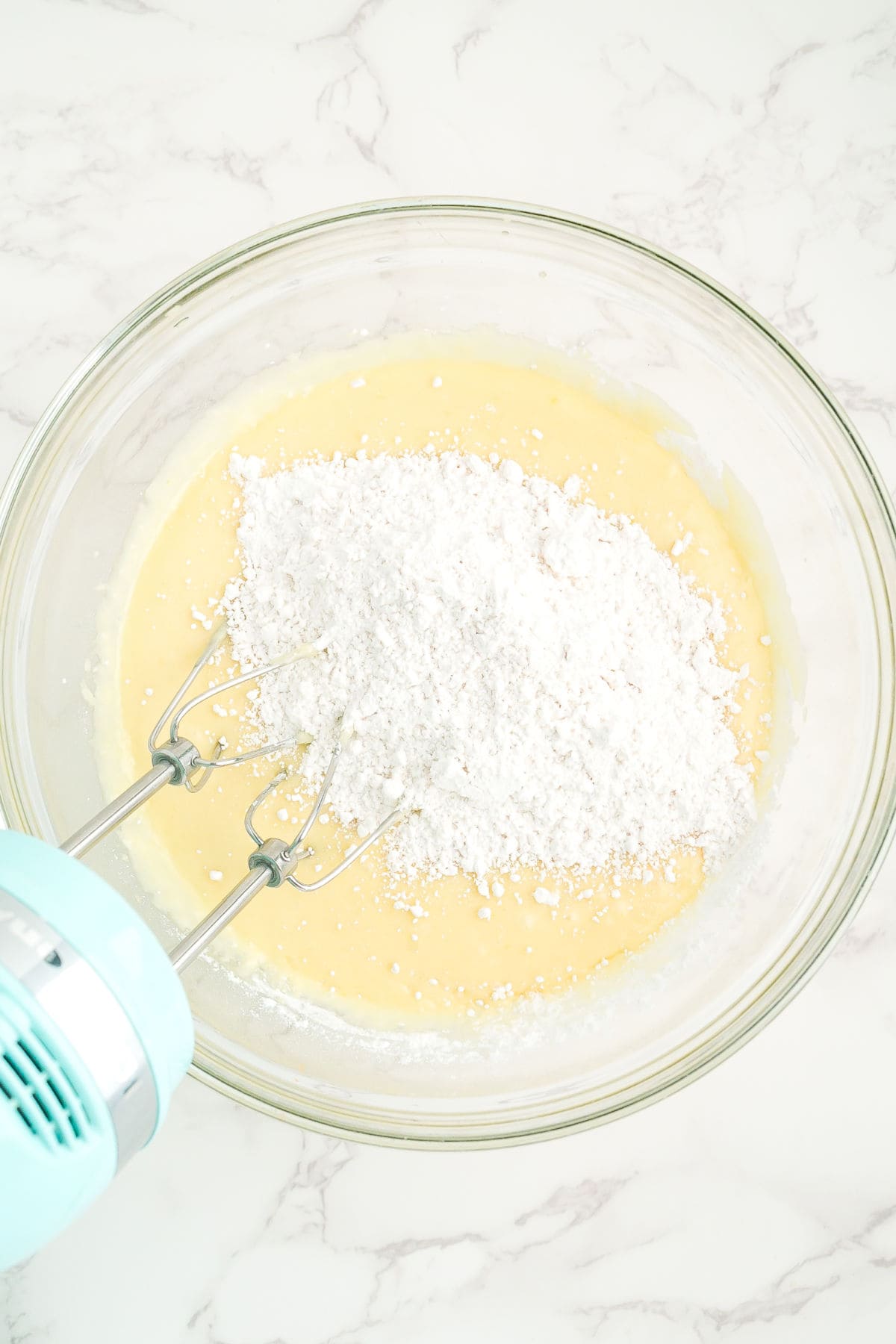 A glass bowl with yellow batter, topped with powdered sugar, being mixed with an electric hand mixer on a marble countertop.