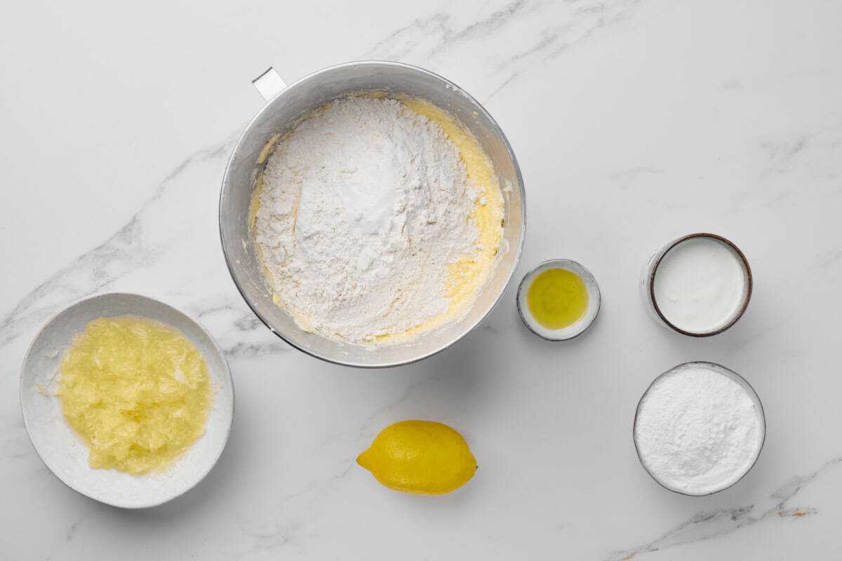 Bowl of flour mixture, plates with lemon zest and powdered sugar, a lemon, a small bowl of oil, and a small bowl of milk on a marble surface.