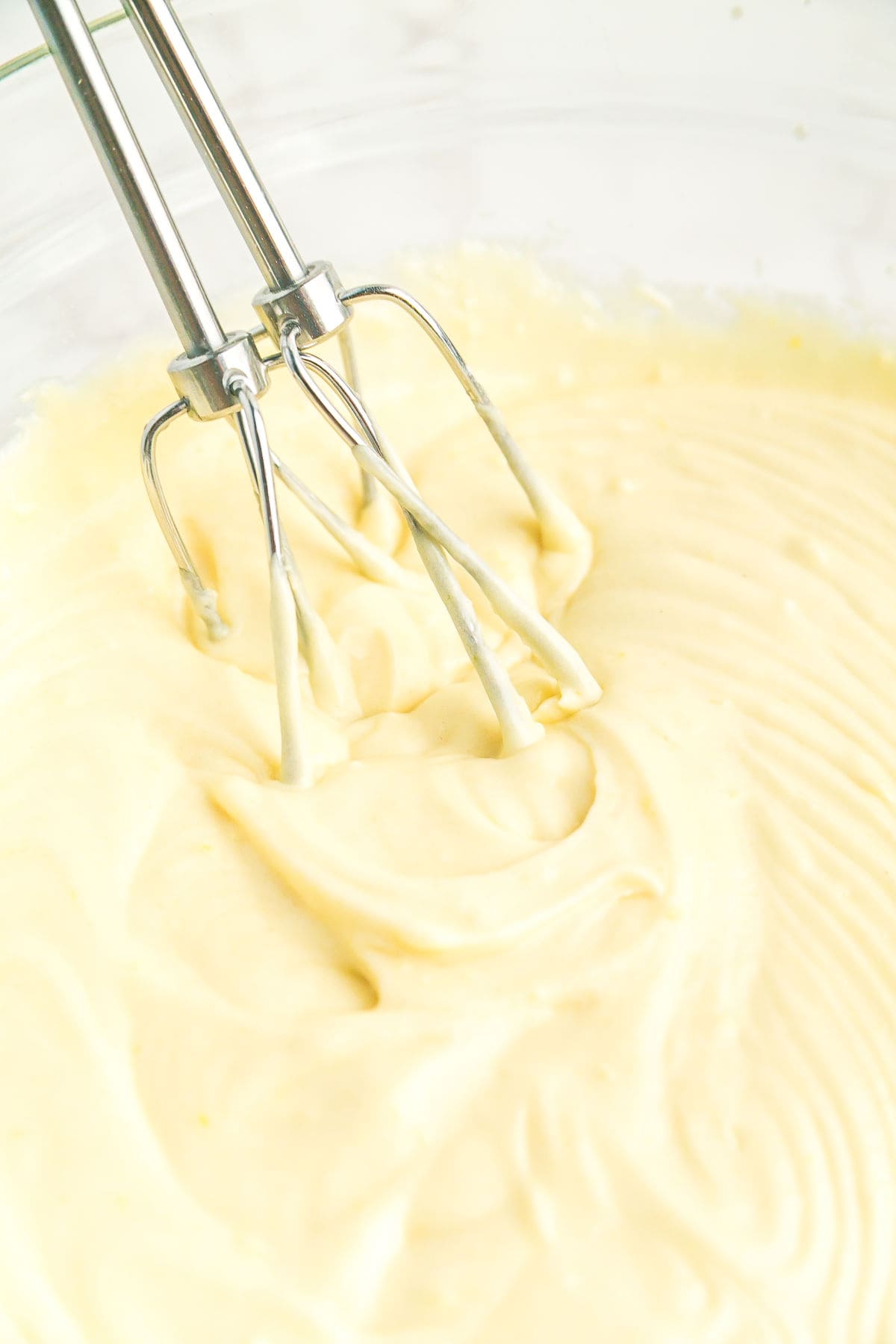 Electric mixer beaters mixing pale yellow batter in a glass bowl.