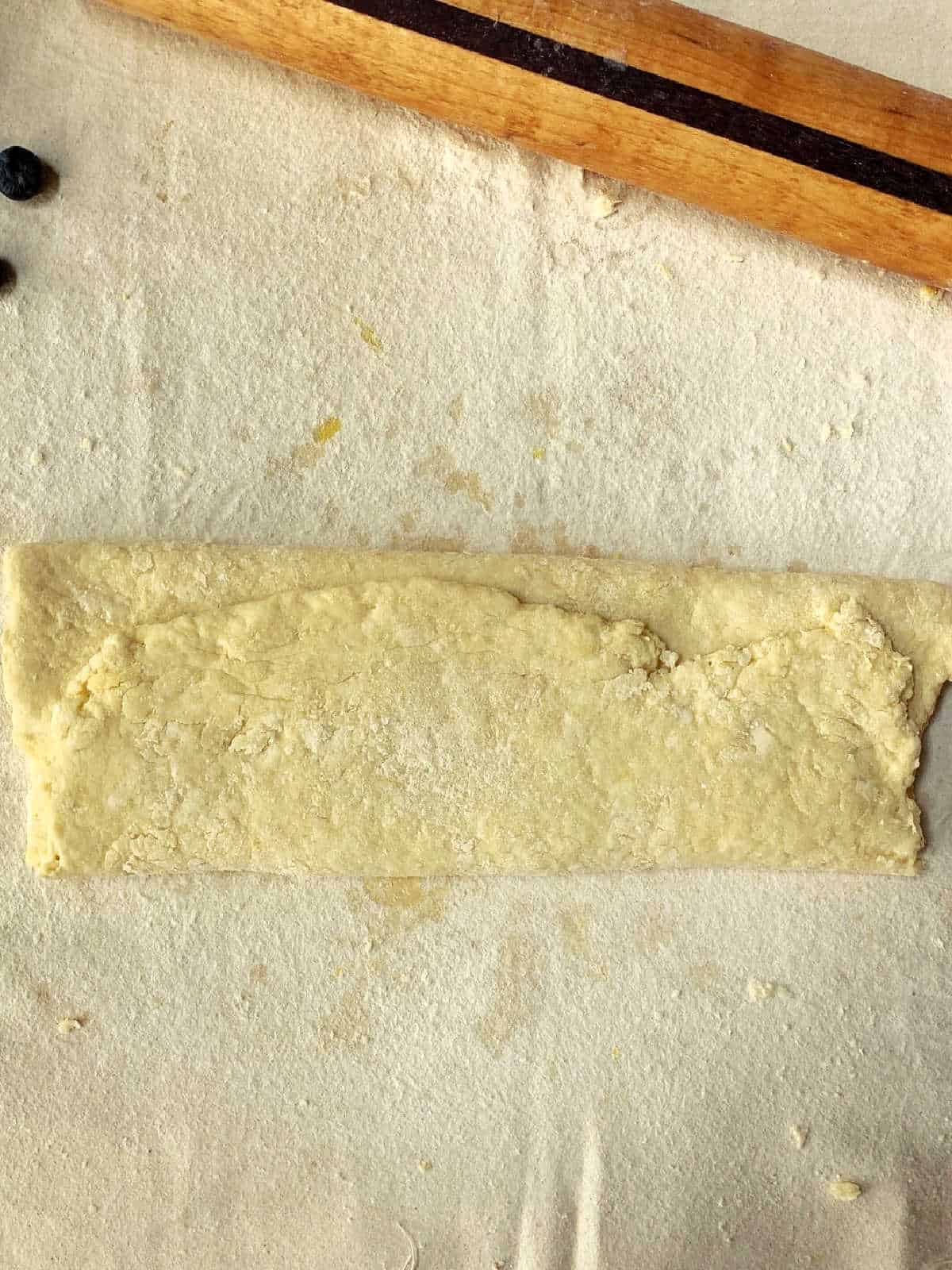 A rectangular piece of folded dough sits on a floured surface with a wooden rolling pin and a few blueberries nearby.