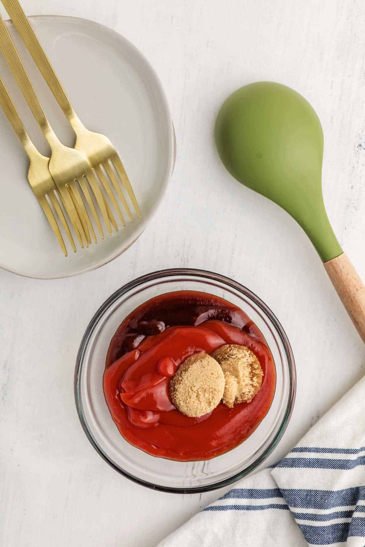 A glass bowl with brown sugar and ketchup on top of a meatloaf mixture, next to a plate with gold forks and a green spatula on a white surface.