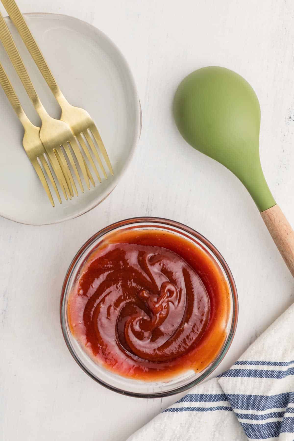 A bowl of barbecue sauce sits on a white surface next to a green-handled spoon, three gold forks on a plate, and a blue-and-white striped cloth.
