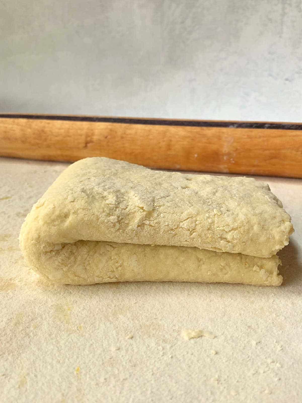 A folded sheet of dough rests on a lightly floured surface with a wooden rolling pin in the background.