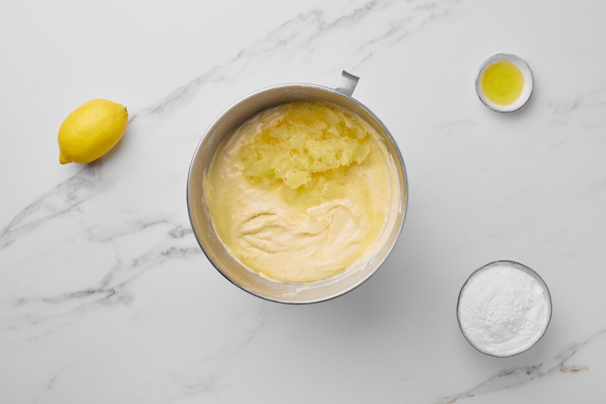 A metal mixing bowl with cake batter and crushed pineapple, next to a lemon, a small dish of oil, and a bowl of flour on a white marble surface.