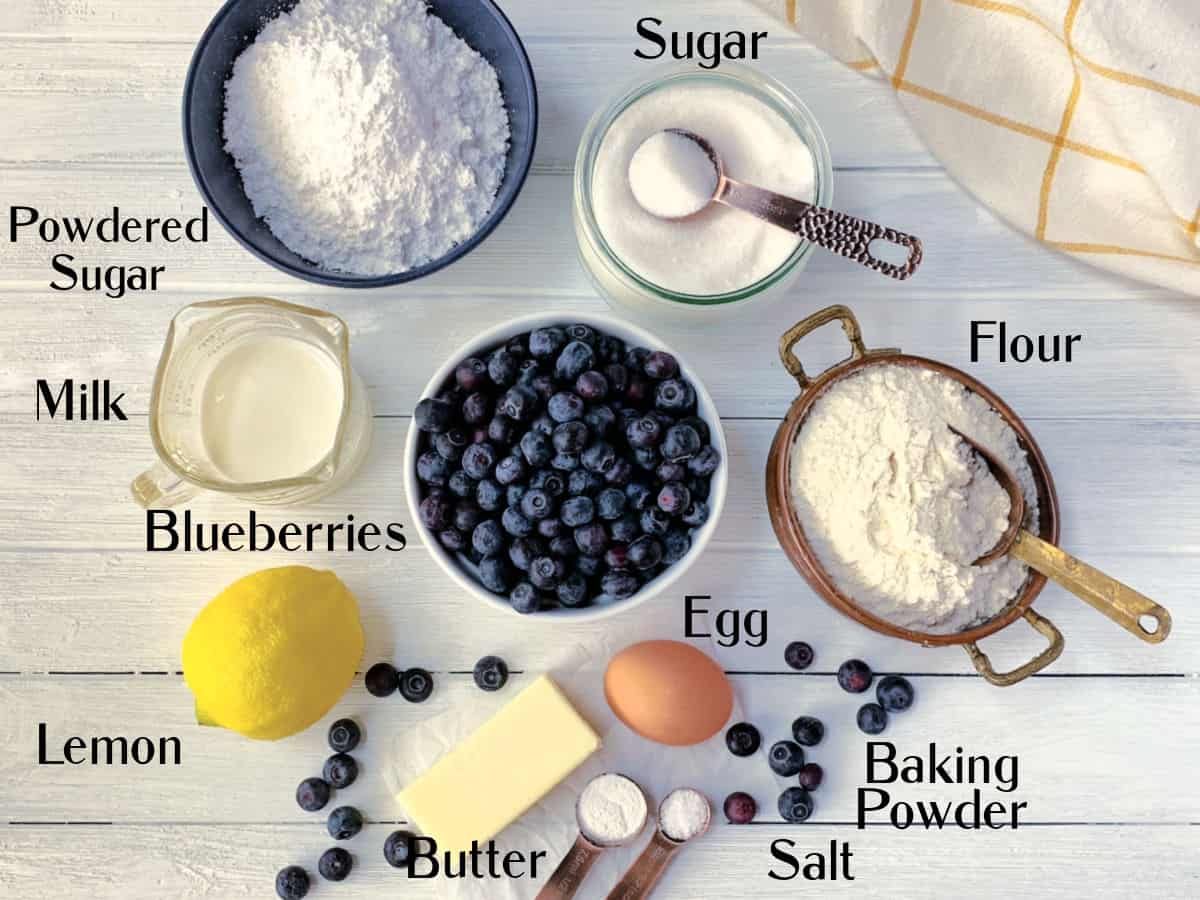 A top view of baking ingredients on a white surface, including blueberries, flour, sugar, powdered sugar, milk, egg, lemon, butter, salt, and baking powder.
