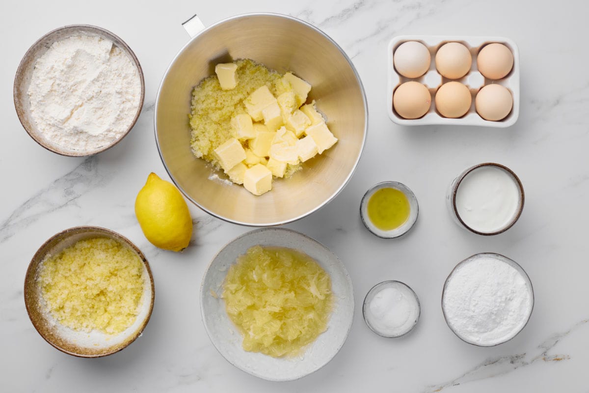 Baking ingredients on a white surface, including flour, sugar, butter, eggs, lemon, lemon zest, lemon juice, yogurt, and powdered sugar arranged in bowls and containers.