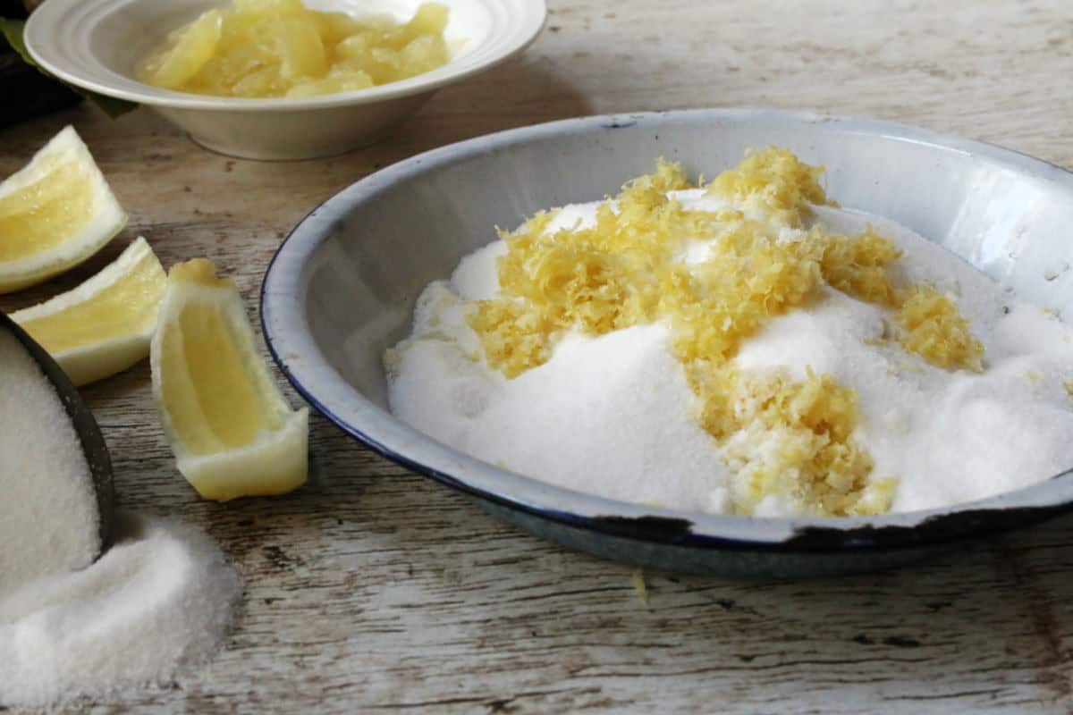 A shallow bowl filled with granulated sugar and lemon zest sits on a wooden surface, with lemon wedges and a bowl of chopped fruit nearby.