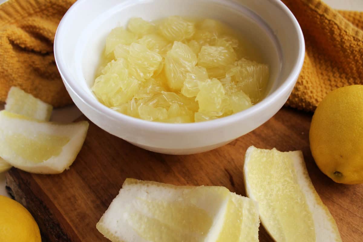A white bowl filled with peeled lemon segments in juice sits on a wooden board, surrounded by pomelo rind, a yellow cloth, and whole lemons.