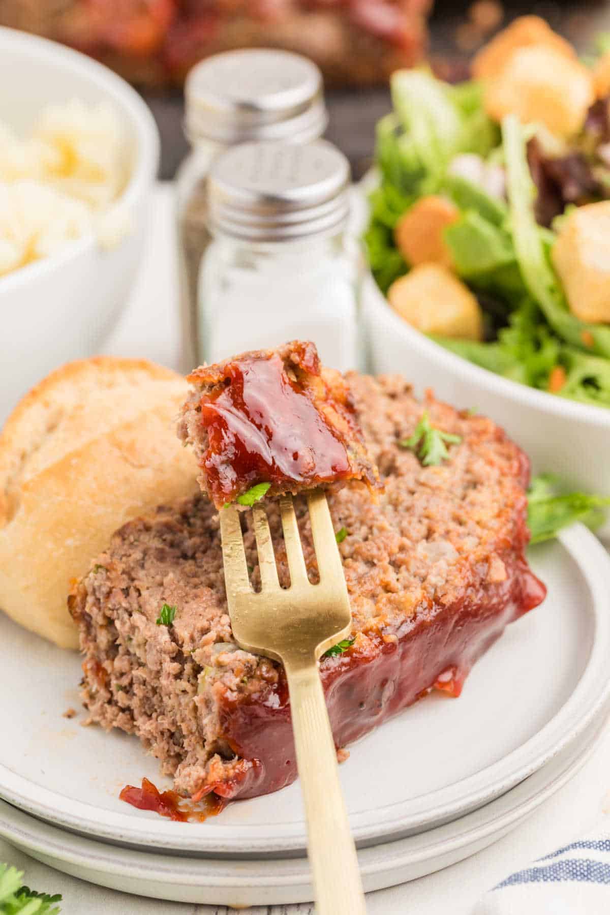 A plate with a slice of glazed meatloaf, a fork holding a bite, a bread roll, mashed potatoes, and a green salad with croutons. Salt and pepper shakers in the background.