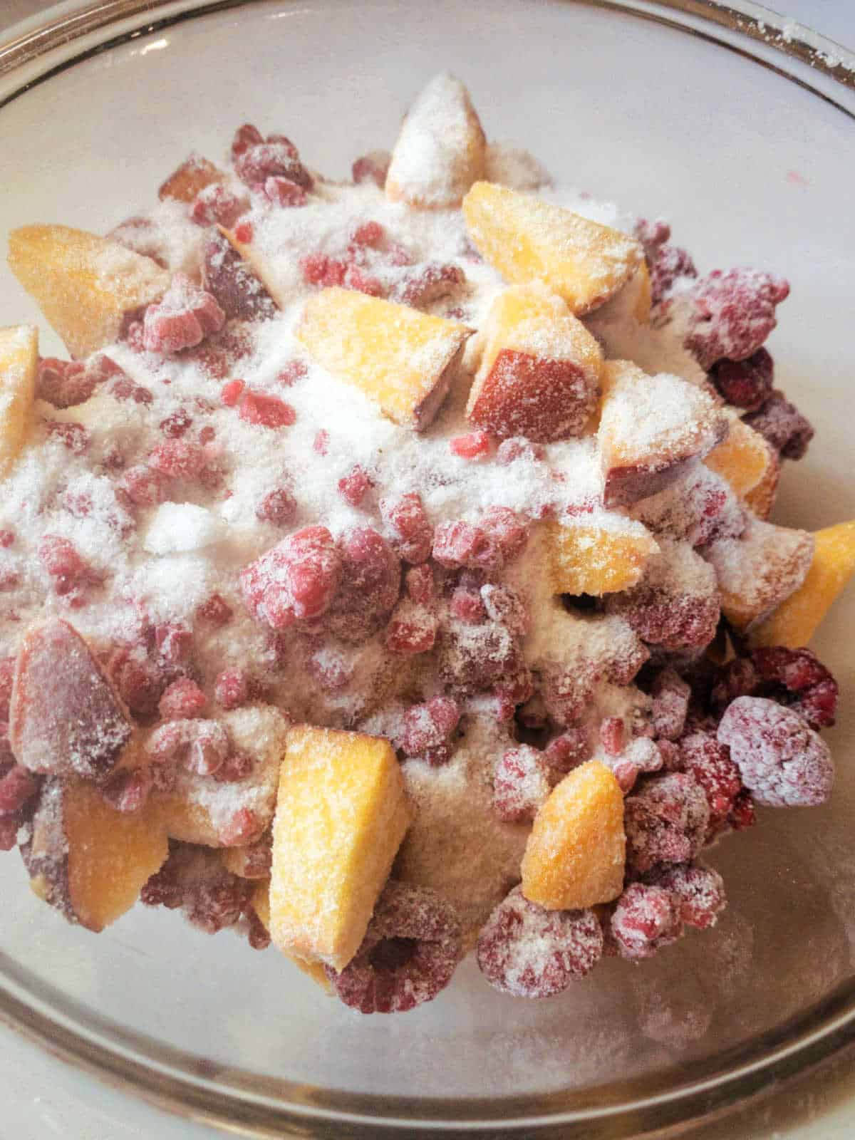 A glass bowl filled with frozen peach slices and raspberries, topped with a layer of granulated sugar.