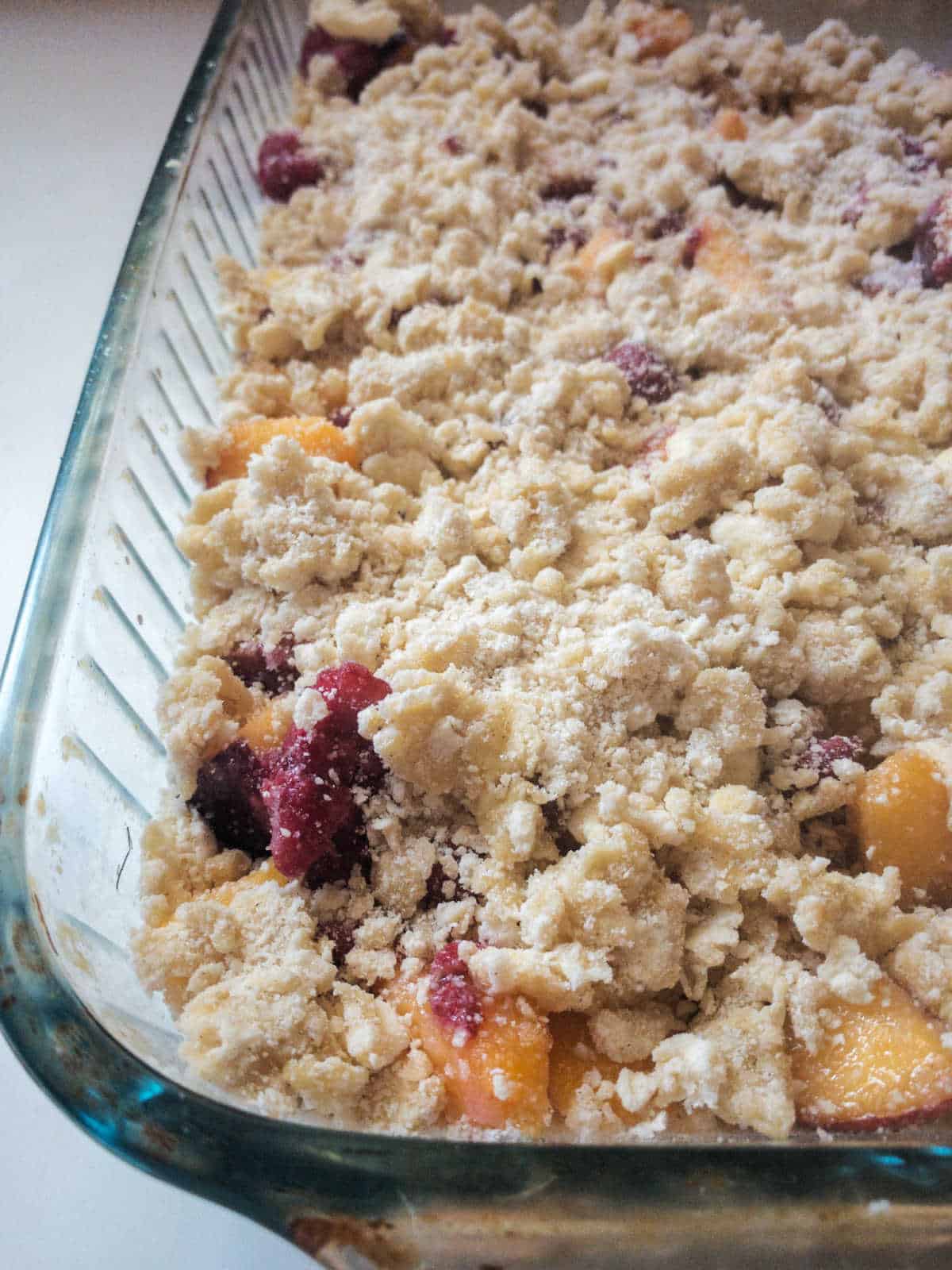 Glass baking dish filled with unbaked fruit crumble, showing pieces of fruit and crumbly topping.