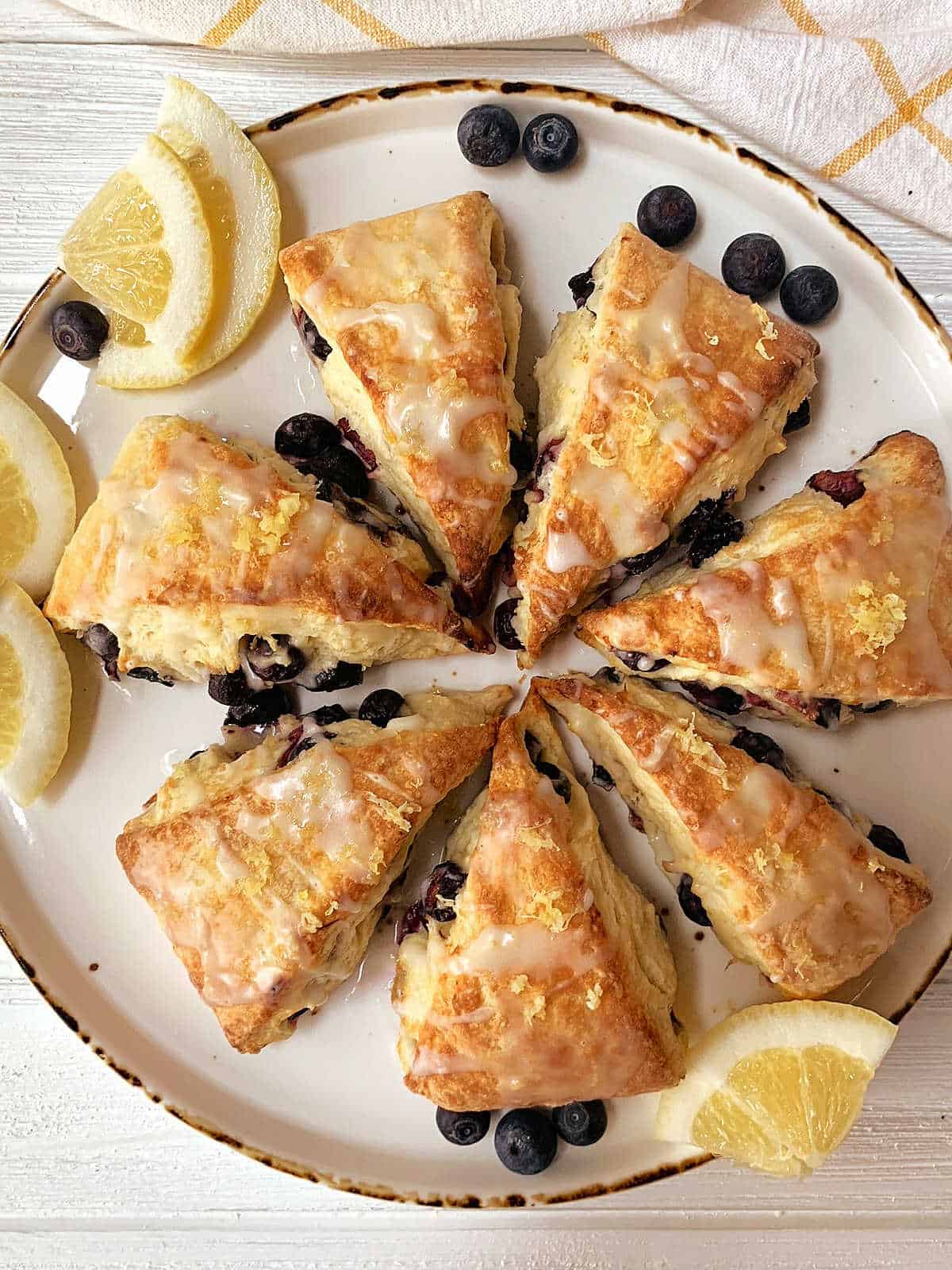 A round plate with eight triangular blueberry lemon scones topped with glaze and lemon zest, garnished with lemon wedges and fresh blueberries.