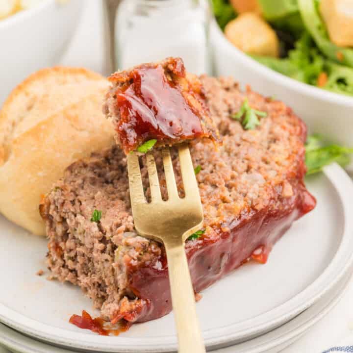 A slice of glazed meatloaf on a plate with a fork, served with a bread roll and salad in the background.
