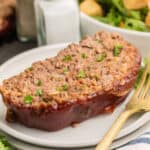 A slice of glazed meatloaf garnished with chopped parsley on a plate with a gold fork, with salad and condiments in the background.