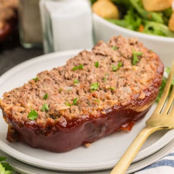 A slice of glazed meatloaf garnished with chopped parsley on a plate with a gold fork, with salad and condiments in the background.
